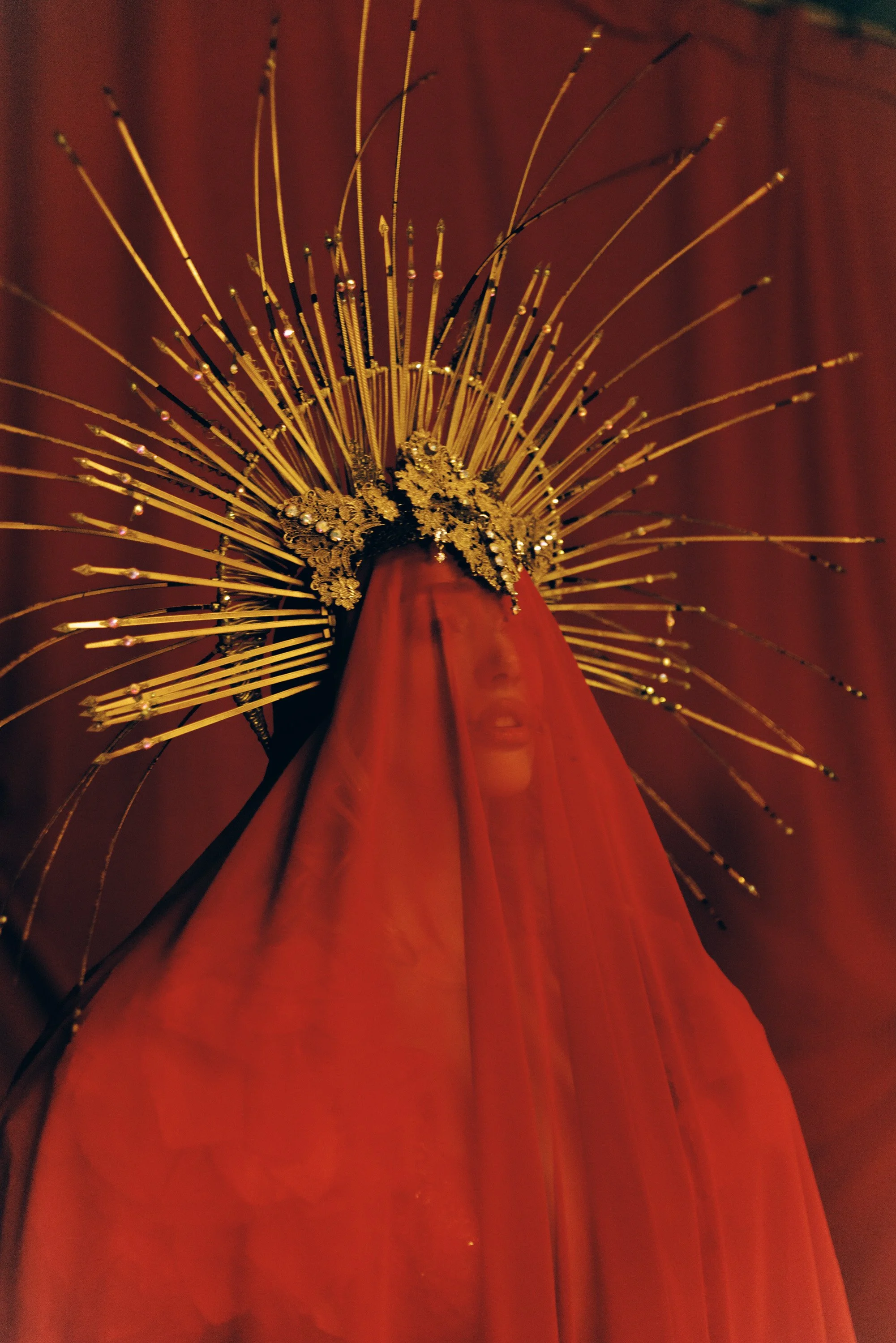 A person dressed as a saint or religious figure, wearing a red robe and an elaborate gold and black halo or headdress with multiple gold rays extending outward, captured from a low angle against a dark background.