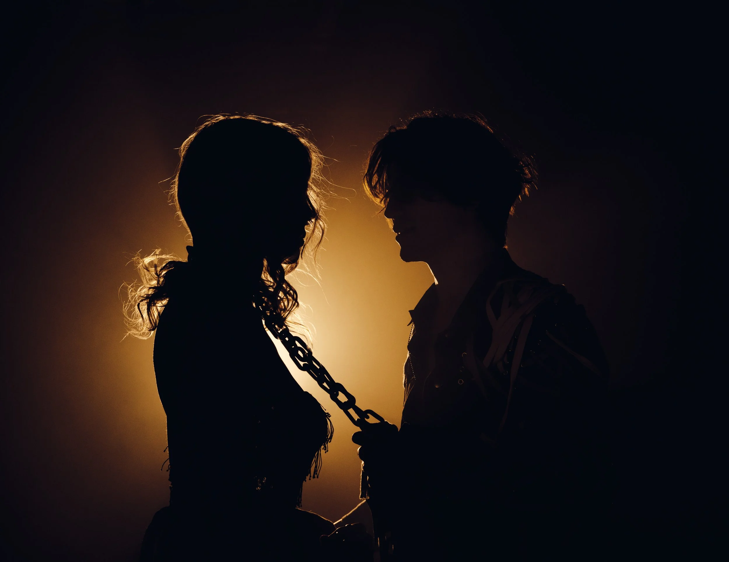 Silhouette of a woman and a man facing each other at night, holding a chain, with warm backlighting highlighting their outlines.
