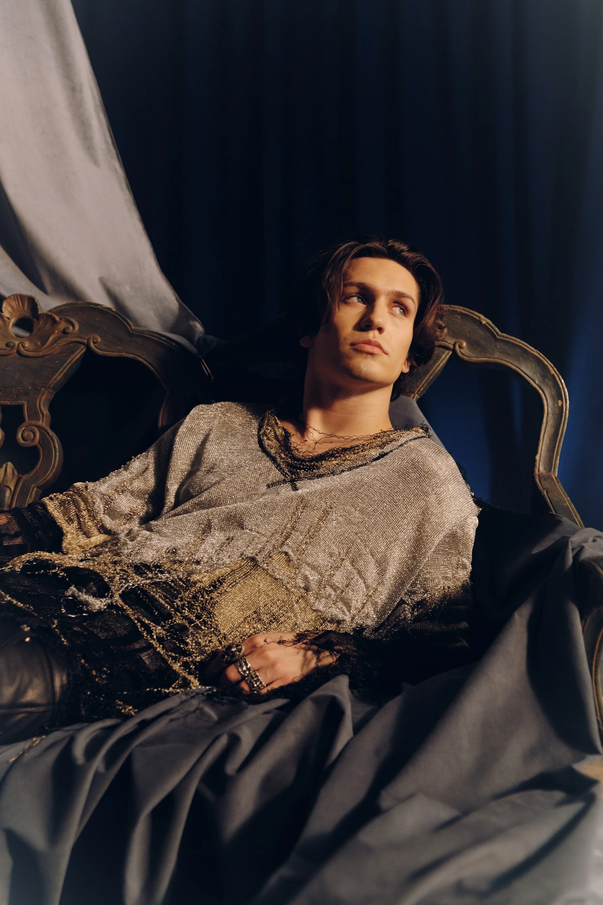 Young man with long dark hair sitting on an ornate, vintage-style couch in a dimly lit room. He is wearing a metallic, shiny top with black lace details and dark trousers, looking contemplative.