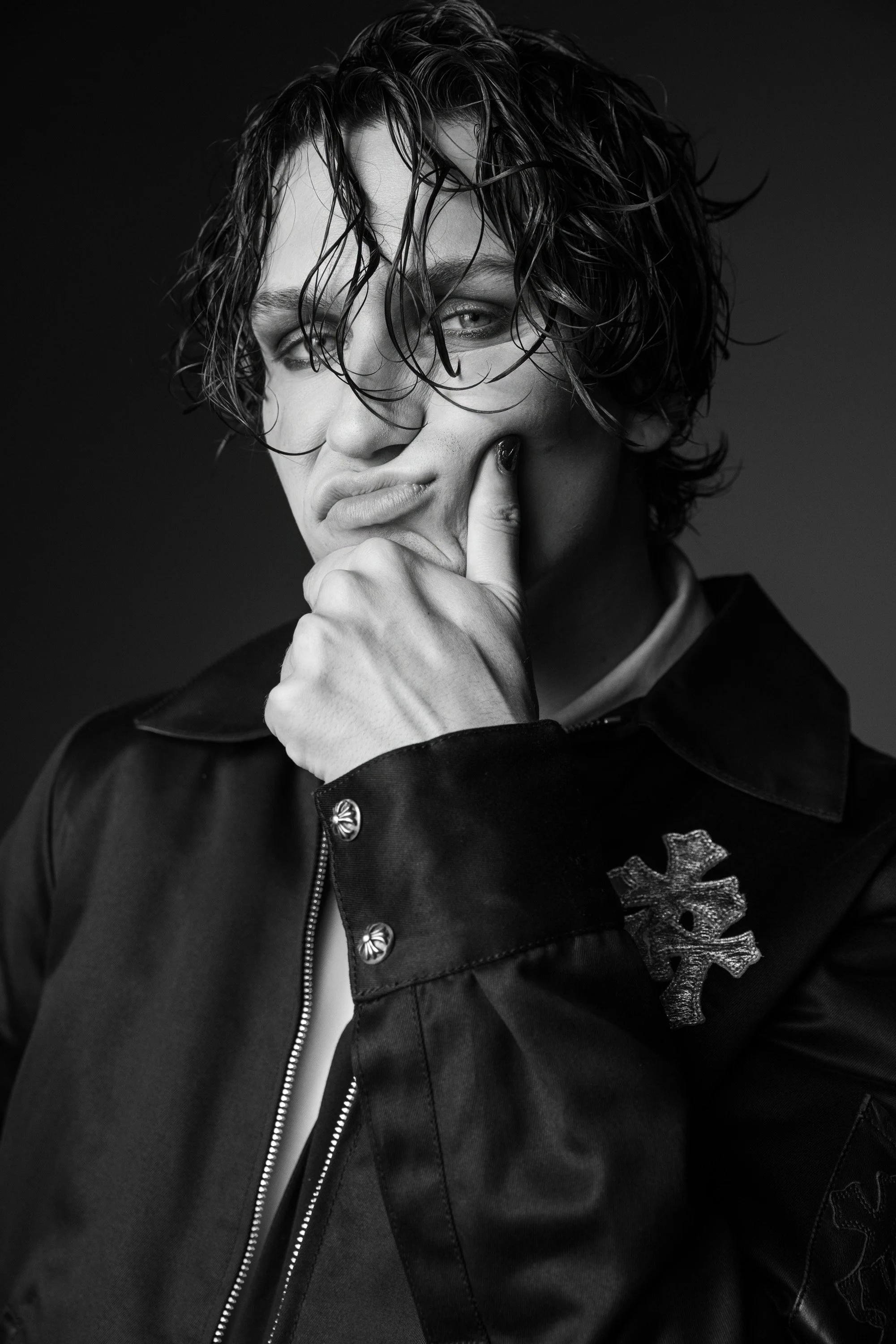 Black and white portrait of a person with wet, curly hair, wearing a dark jacket with buttons and a patch of the skull and crossbones design, touching their chin with their hand and looking at the camera.