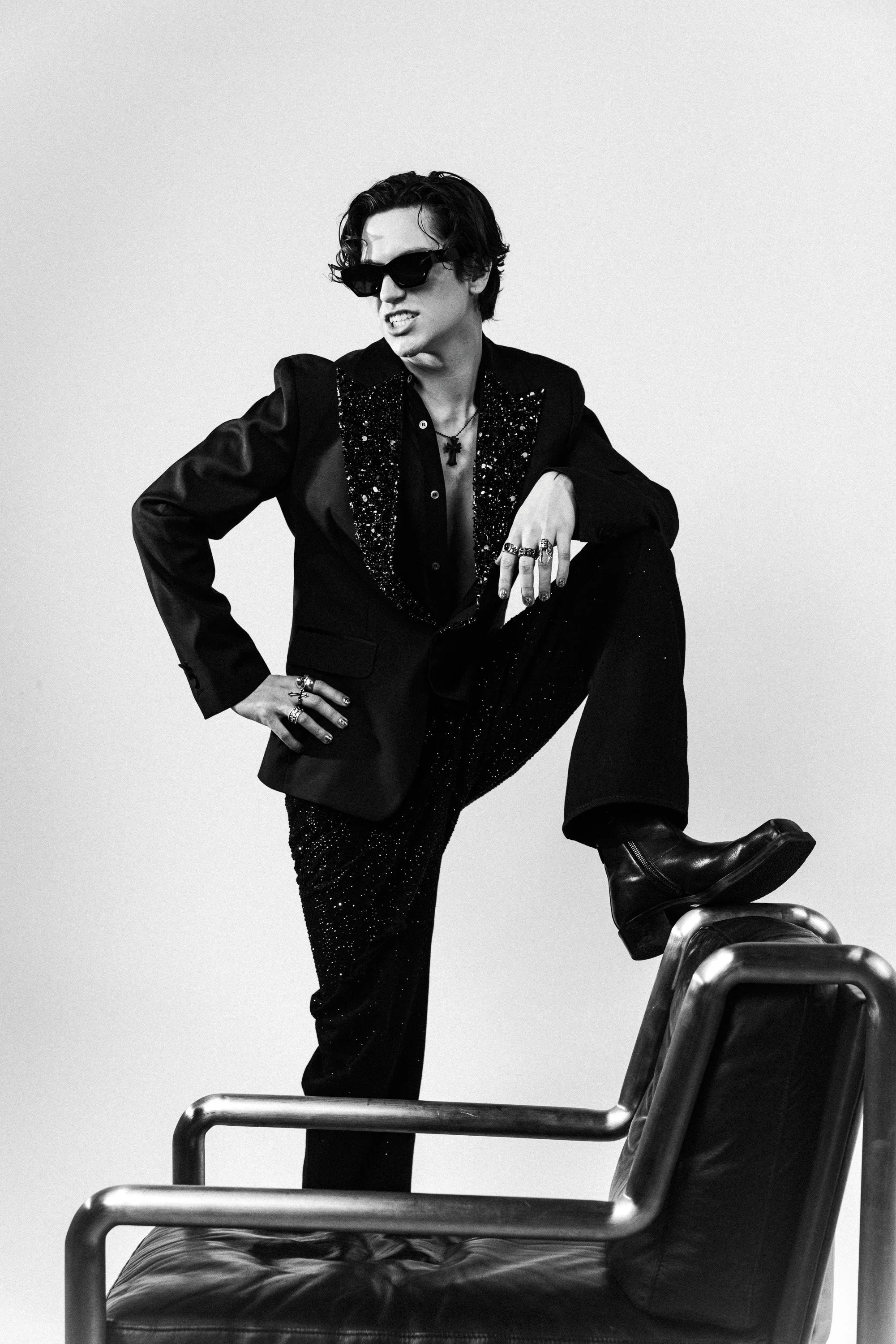 Black and white photo of a person wearing sunglasses, a black blazer with glittery details, and multiple rings. They have dark wavy hair and a confident expression, striking a pose with one leg raised on a chair and one hand on their hip.