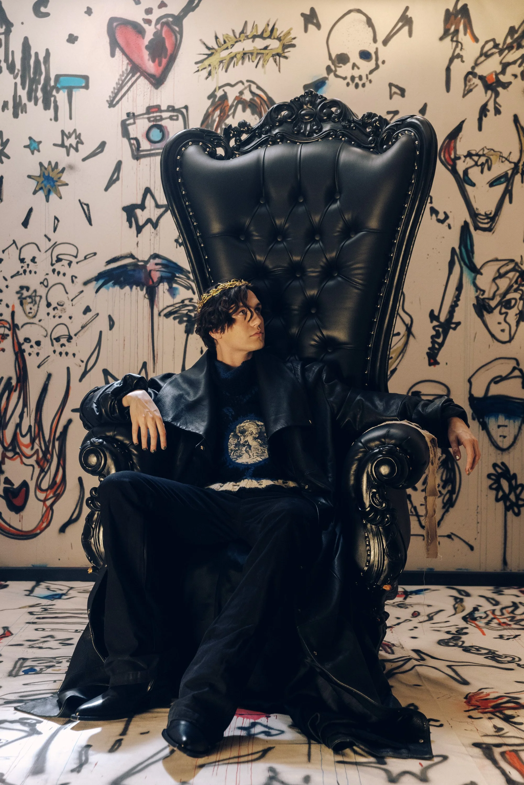 A person with dark, wavy hair sitting in a large ornate black throne with a high backrest, against a graffiti-style wall featuring various drawings of faces, skulls, hearts, a crown, and other symbols.