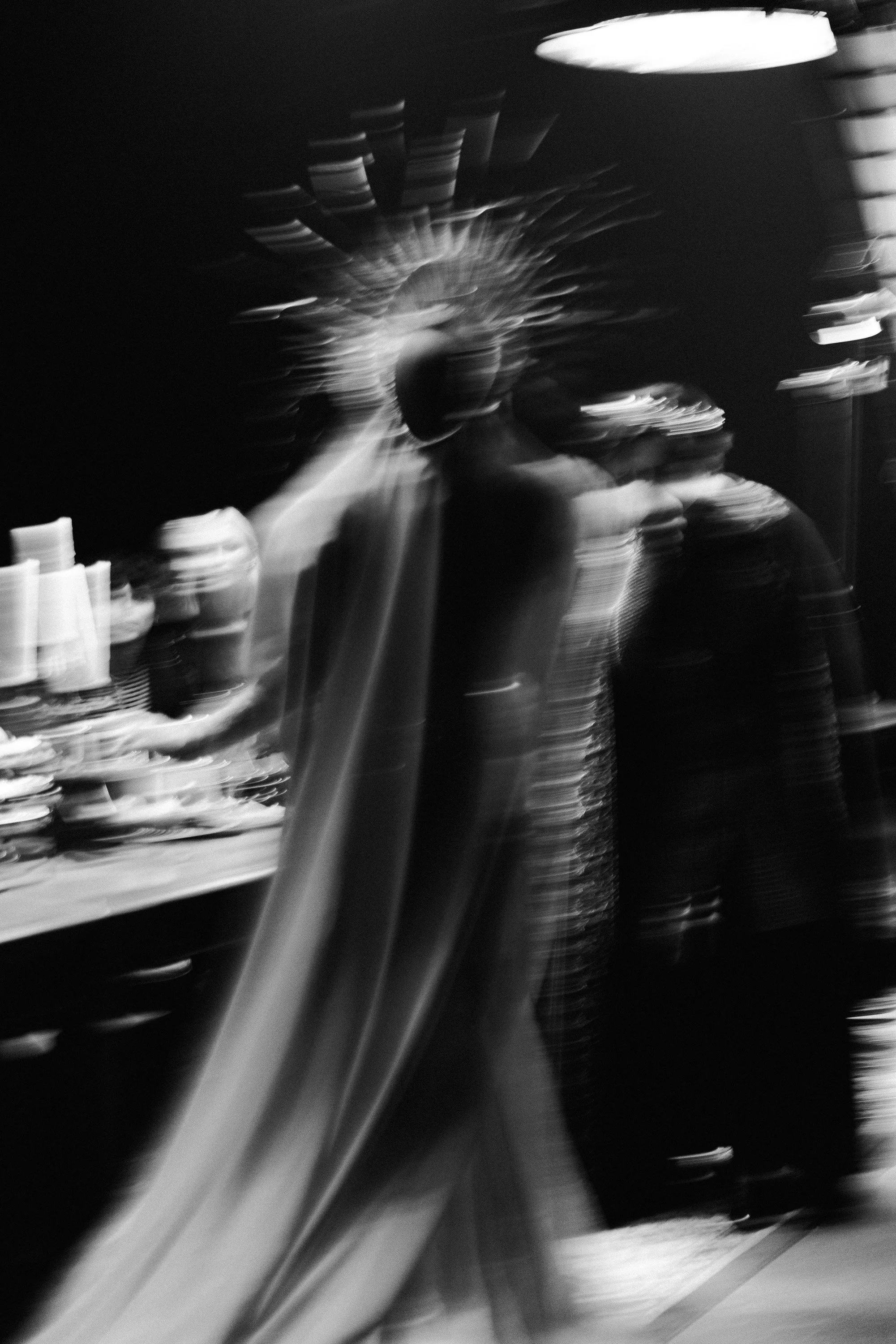 Blurry black-and-white photograph of a person standing behind a table with stacked cups or containers, with abstract light streaks and shapes in the background.