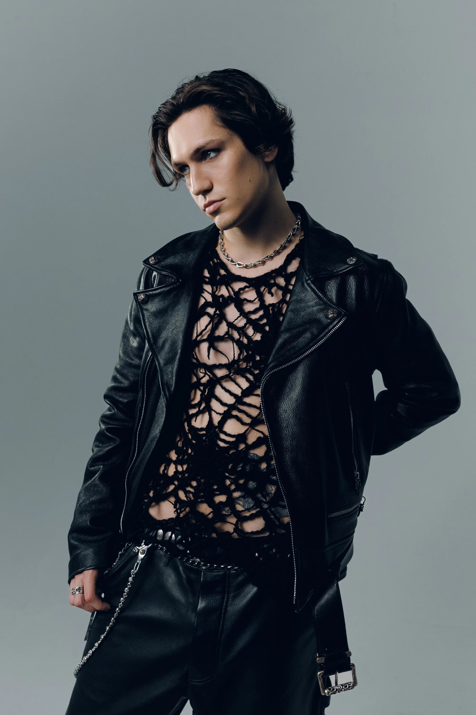 A young person with dark, wavy hair posing against a plain gray background. They are wearing a black leather jacket over a see-through black lace top, accessorized with layered silver necklaces and rings, and holding a small black purse with a chain 