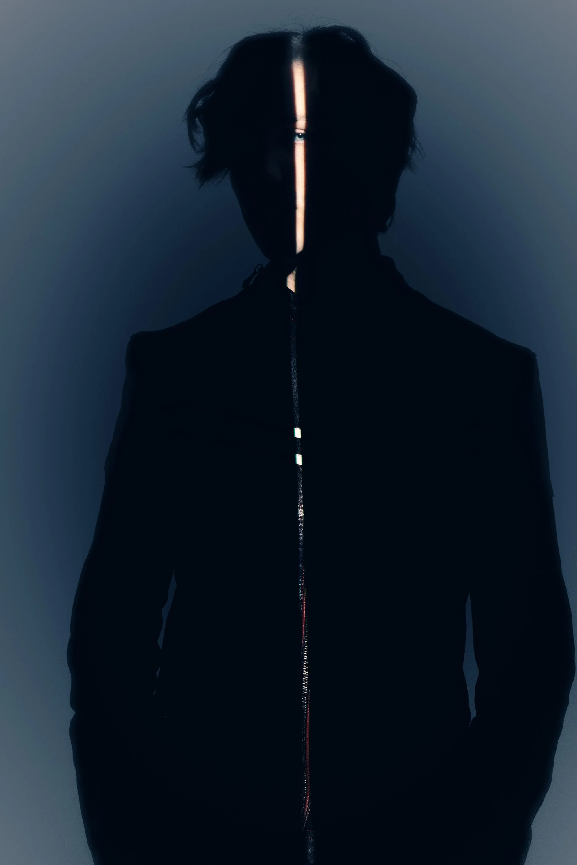 A person with black hair, wearing a black jacket with a zipper, standing in front of a dark background with a vertical light reflection on their face and chest.