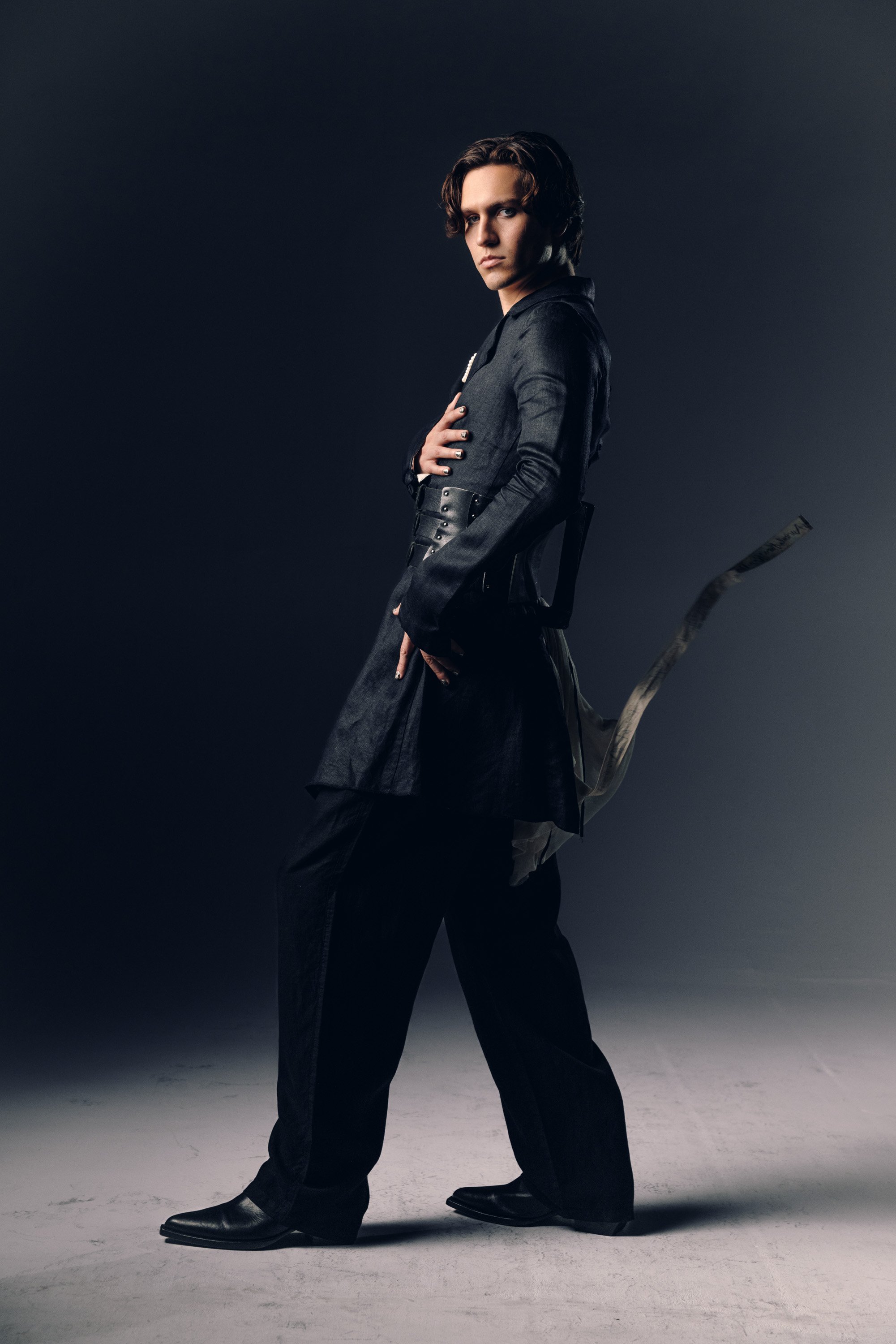 A young man with brown, wavy hair, dressed in a dark, stylish outfit, stands against a dark background. He is looking directly at the camera with a serious expression and has one hand on his chest. His look is edgy and fashion-forward, with pointed s