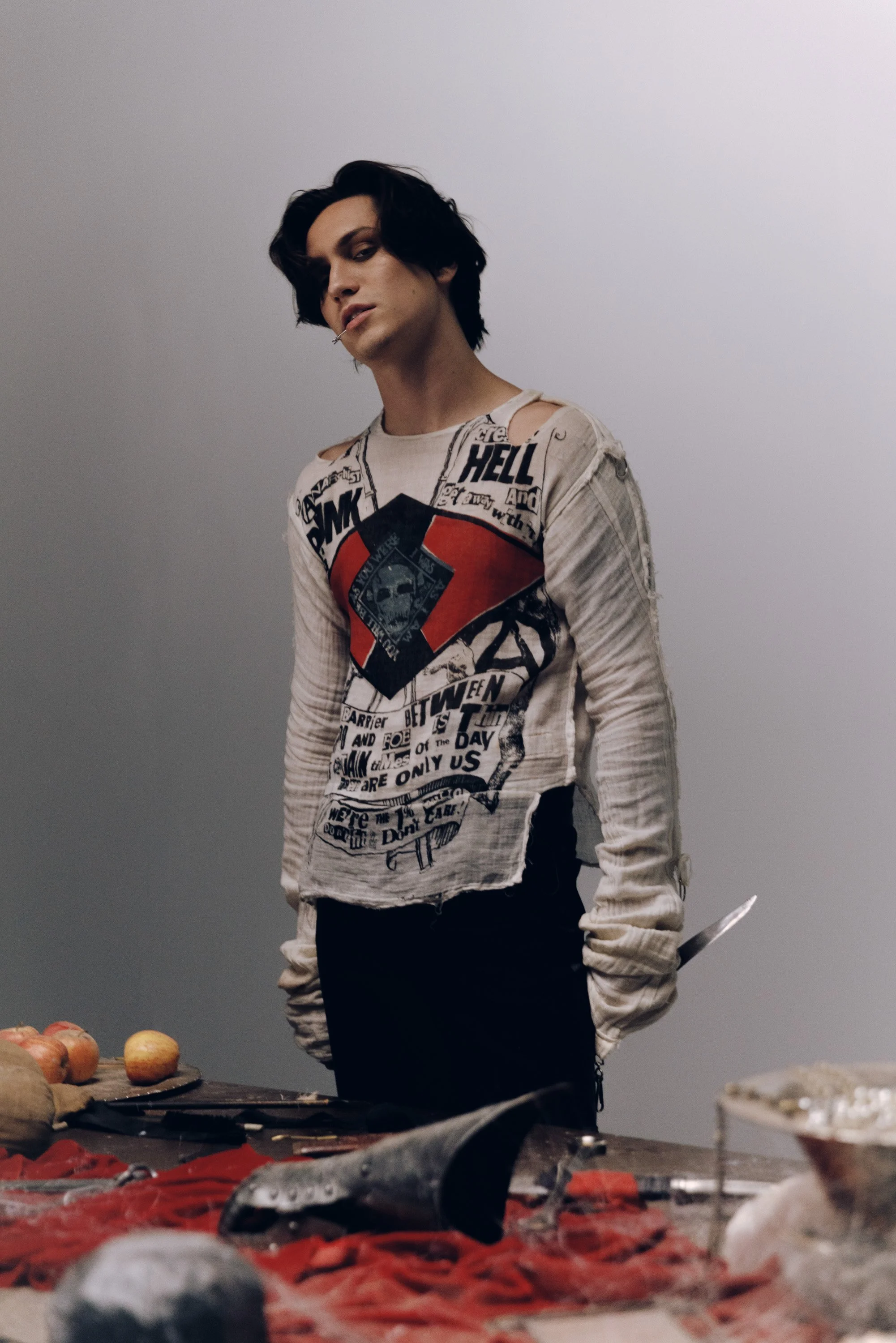 A young man with black hair and a cigarette in his mouth standing in front of a table with apples and a large knife, wearing a distressed graphic shirt with text and symbols, in a minimal indoor setting.