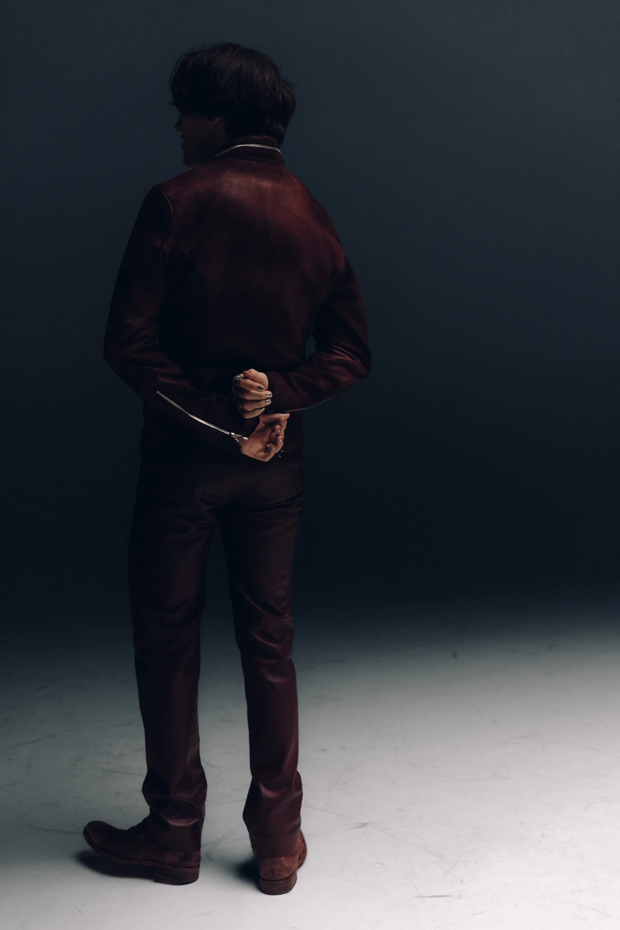 A person standing with their back turned, wearing a dark red leather jacket and matching pants, with their hands clasped behind them, against a dark background.