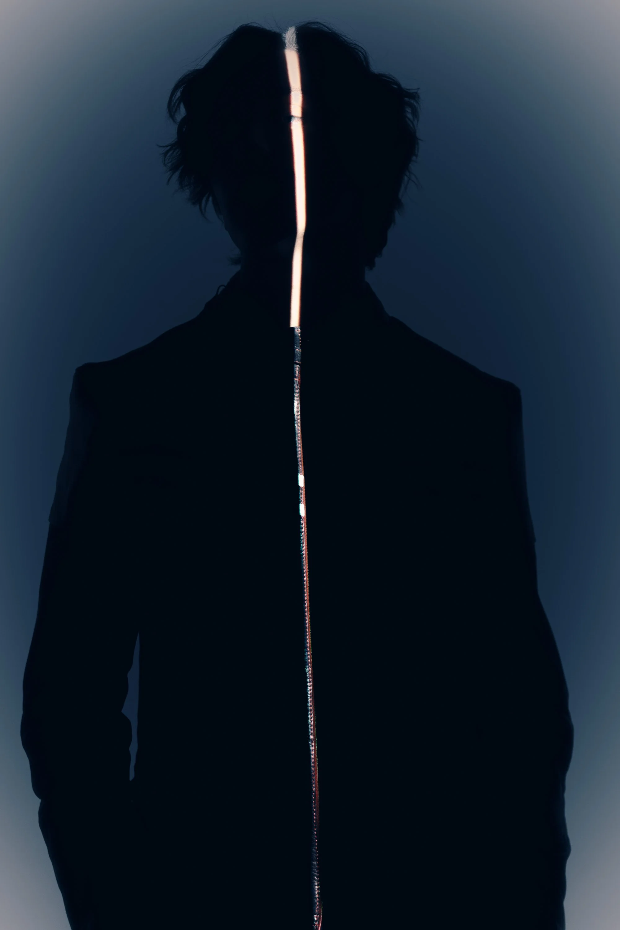 Silhouette of a person with dark, messy hair, standing against a dark background, illuminated by a bright, vertical light strip down the middle of their face and chest.