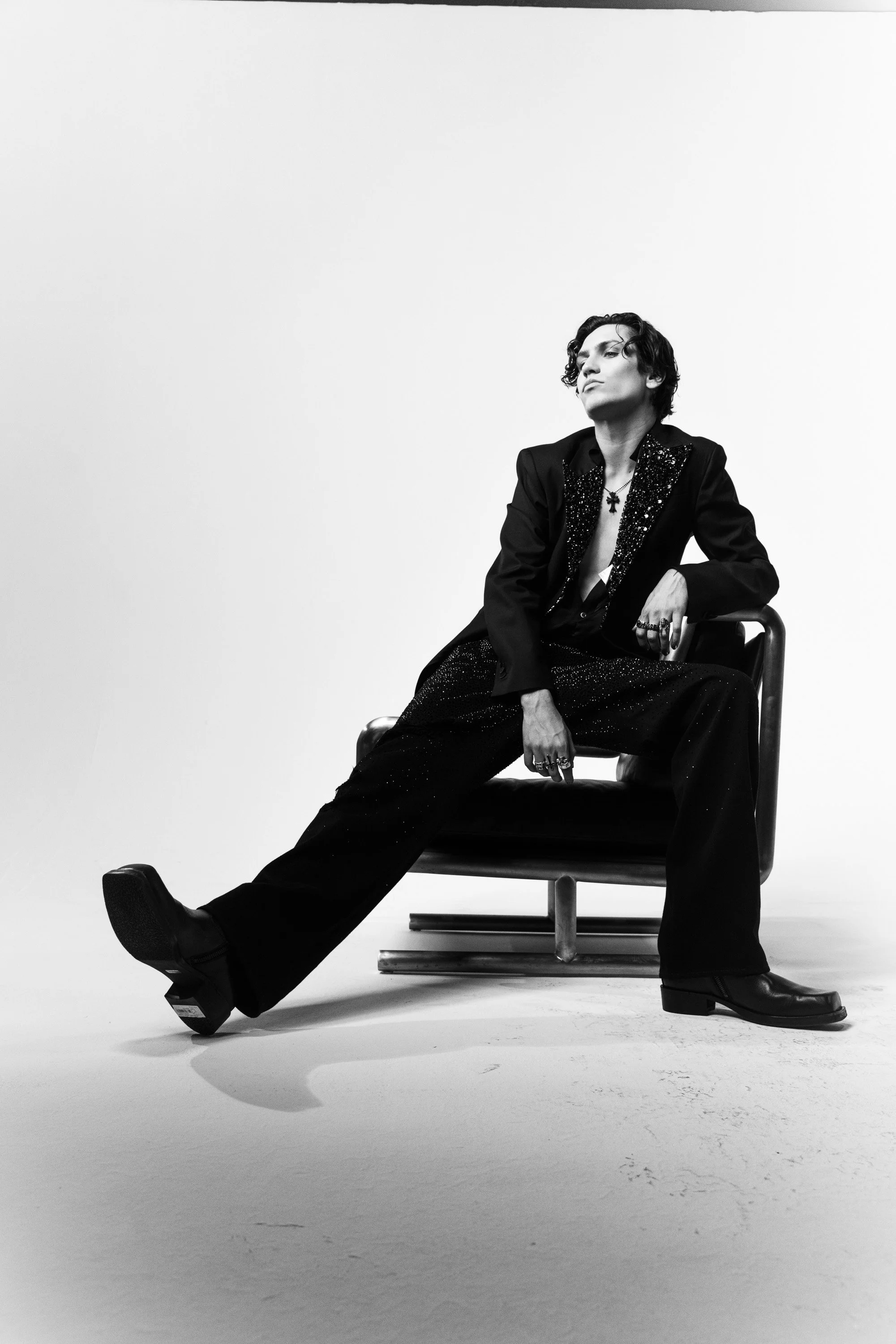 Black and white photo of a person with curly hair sitting on a chair, wearing a black blazer with sequins, loose fitting pants, and black boots, posing with one arm on the chair and the other on their knee.