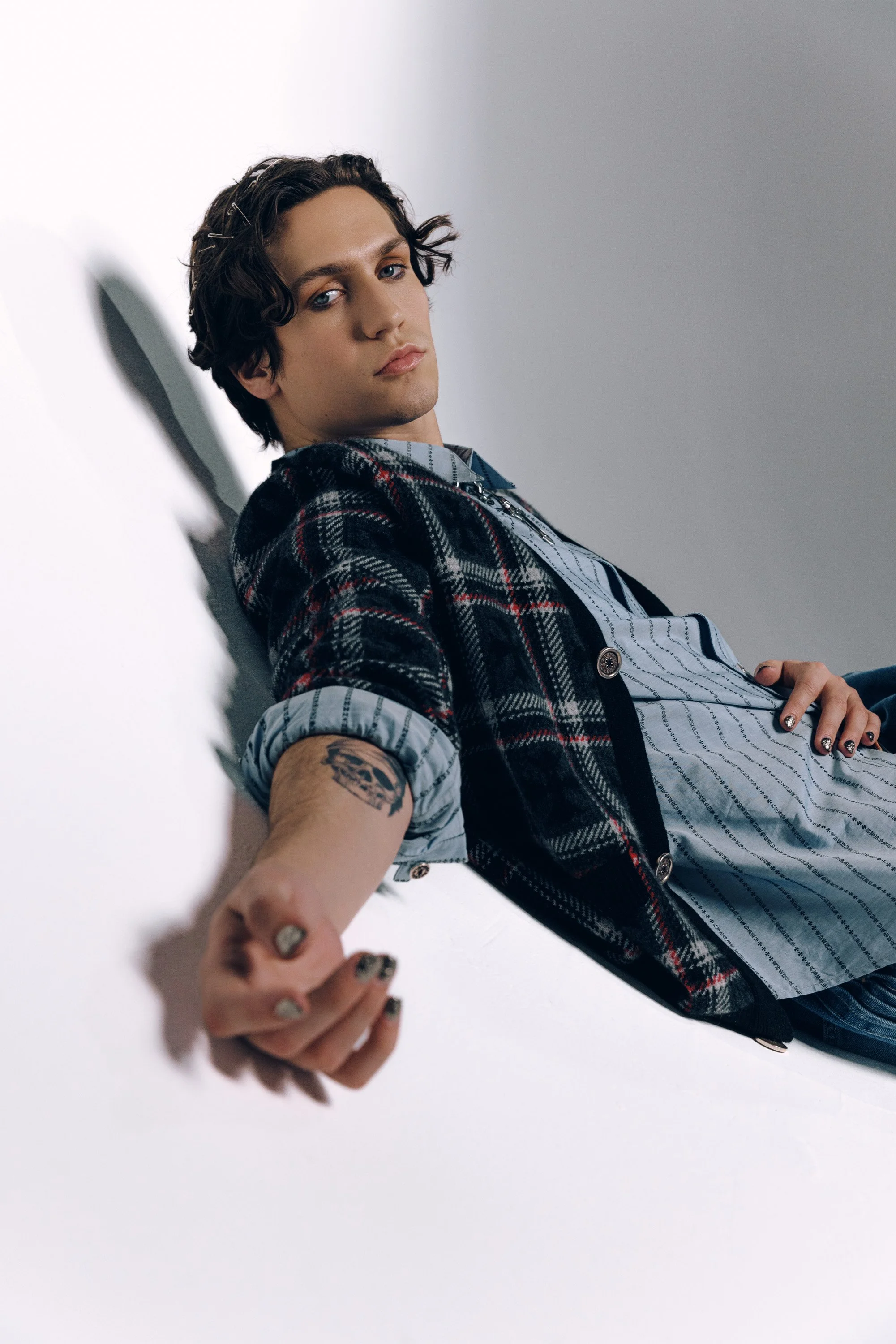 Young man with dark curly hair, blue eyes, and tattoos on his arm, leaning against a white wall, wearing a plaid blazer over a striped shirt.