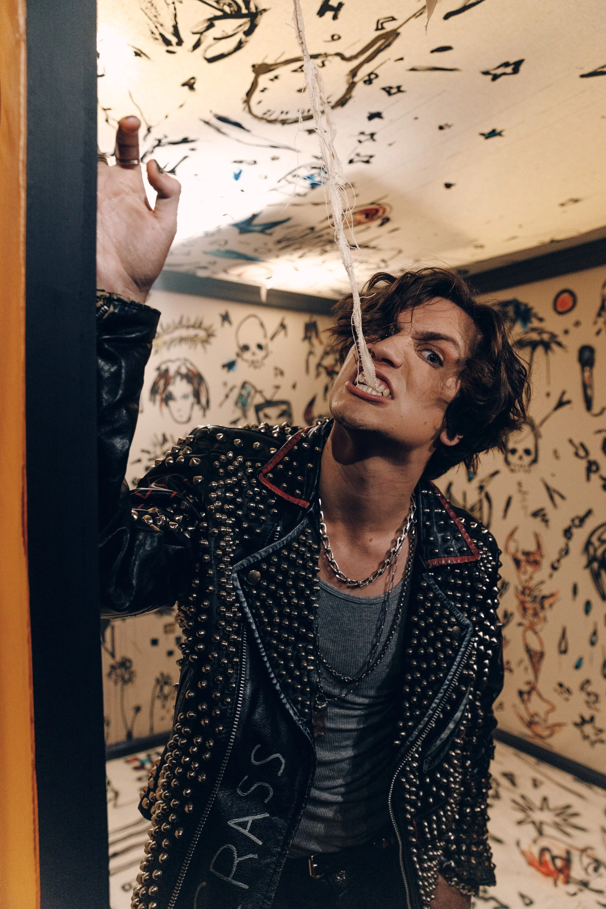 A young man with curly hair wearing a black studded leather jacket and layered necklaces is posing indoors in front of a wall covered with tattoo-style art and skull illustrations. He is making an exaggerated facial expression with his tongue out and