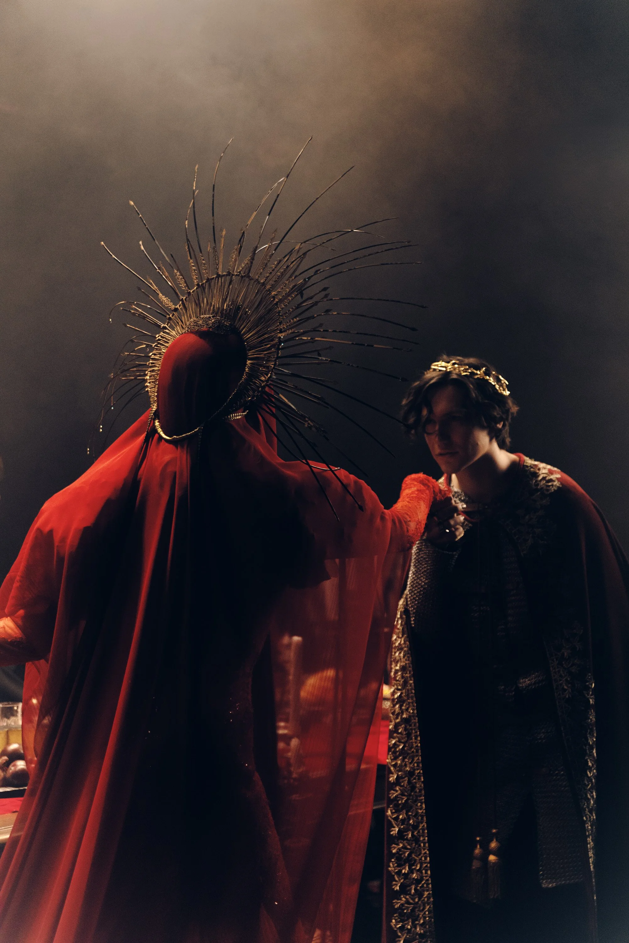 A person in ornate, gold-accented clothing, with short dark hair and glasses, appears to be assisting a figure dressed in a large, dark red costume with an intricate, spiked headpiece, in a dark, possibly indoor setting.