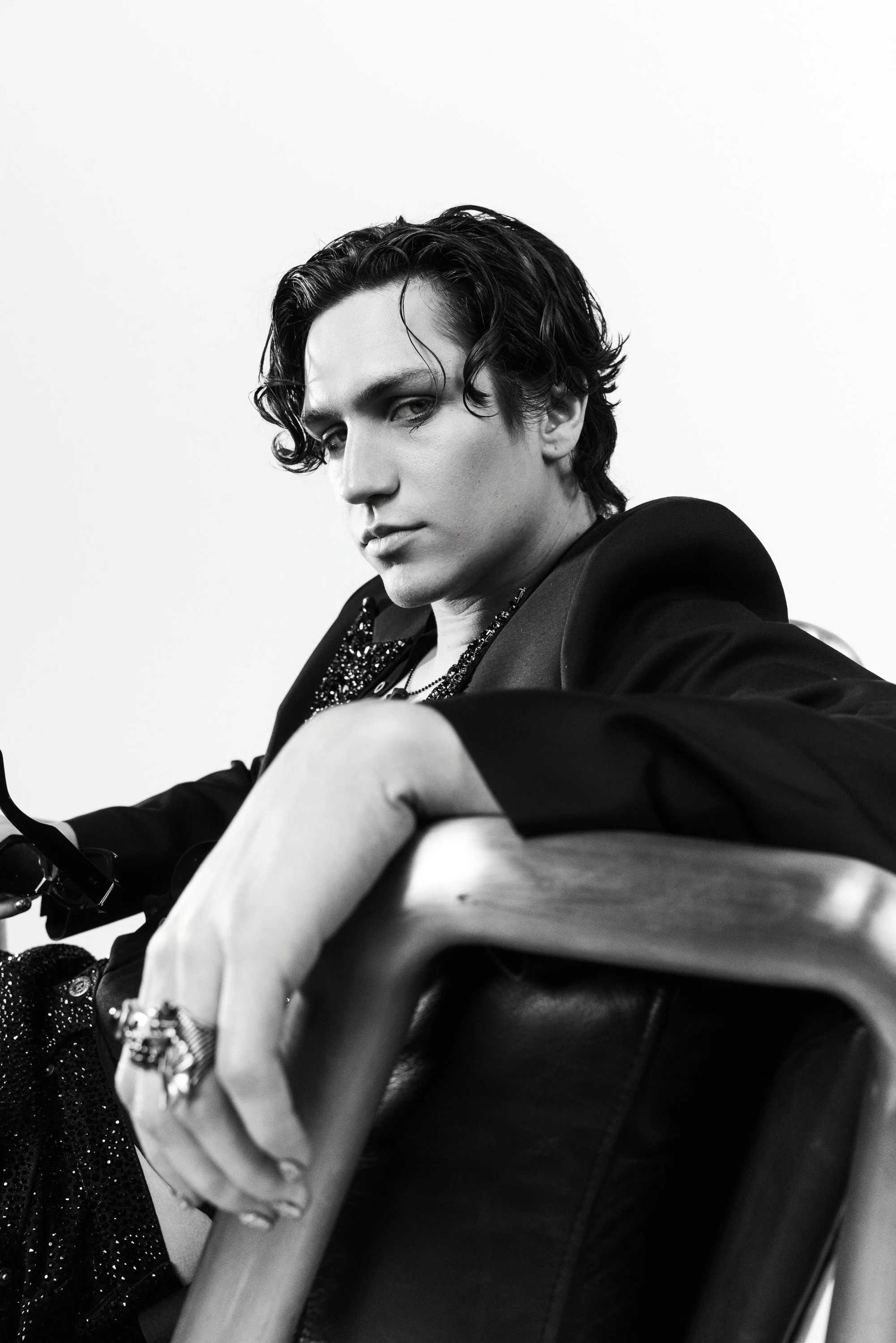 Black and white portrait of a young man with wavy dark hair, wearing a blazer and jewelry, sitting with his arm resting on a chair, looking at the camera.