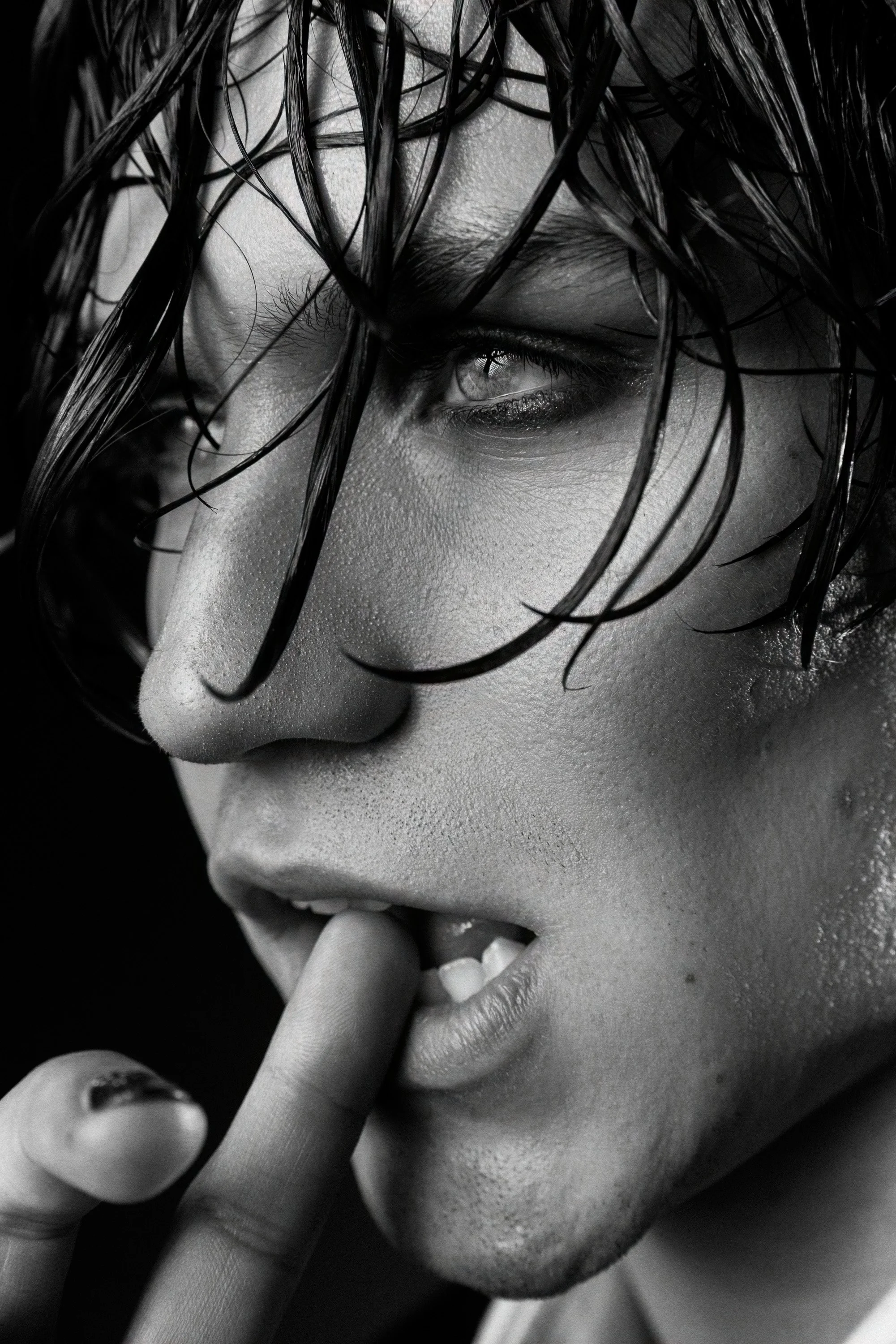 Close-up black and white photo of a woman's face with wet hair partially covering her eye, her finger touching her lips, and detailed skin texture visible.
