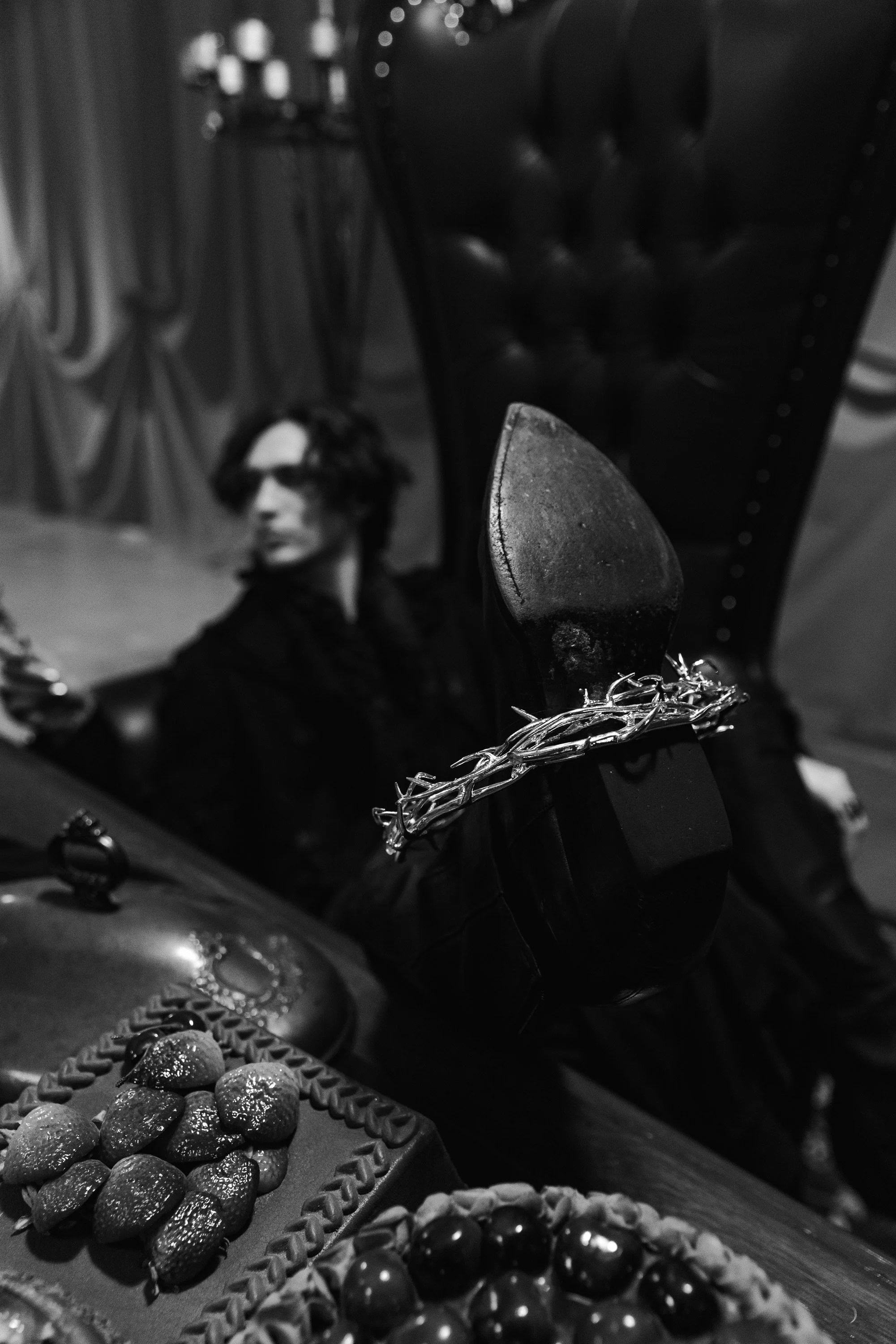 Black and white photo of a stuffed mannequin in the background with short dark hair, wearing glasses and a dark shirt. In the foreground, there is a shoe with a high heel resting on a surface, with jewelry including a chain and rings close by. There 