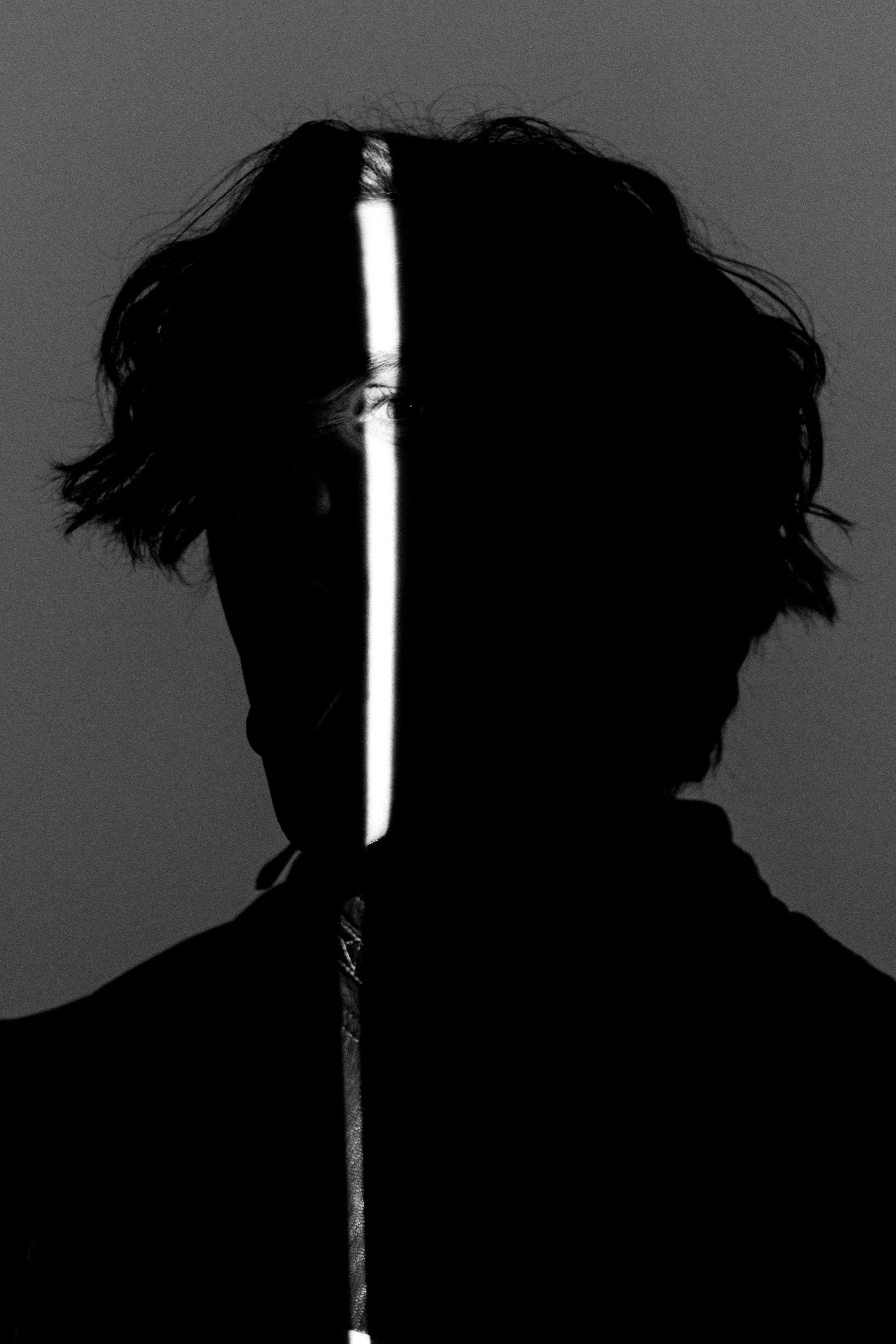 Silhouette of a person with disheveled hair, facing the camera, with a bright vertical light dividing their face into two halves, against a dark background.
