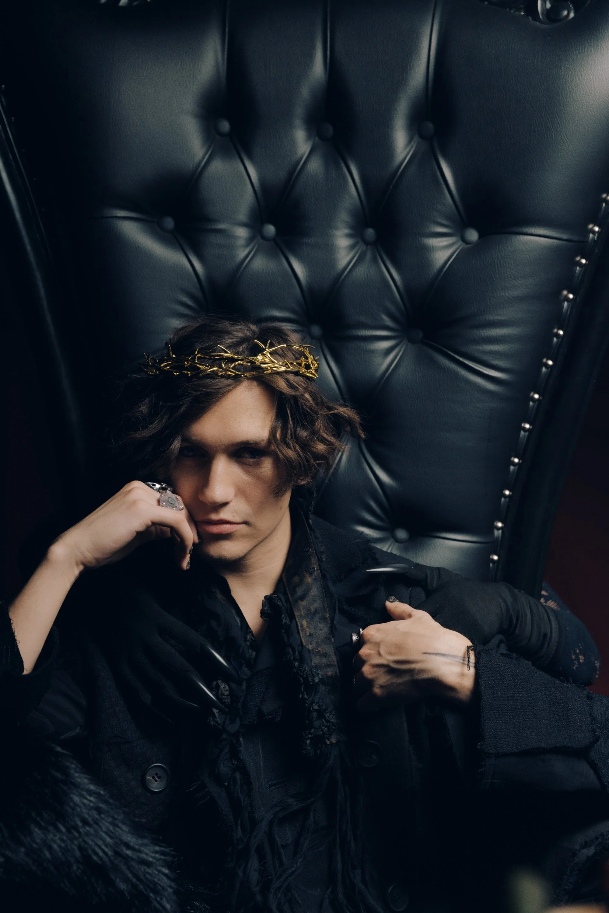 Person with wavy hair and a gold crown sitting on a black leather tufted throne, wearing a black leather jacket, with a silver ring on their finger, holding a finger near their face.