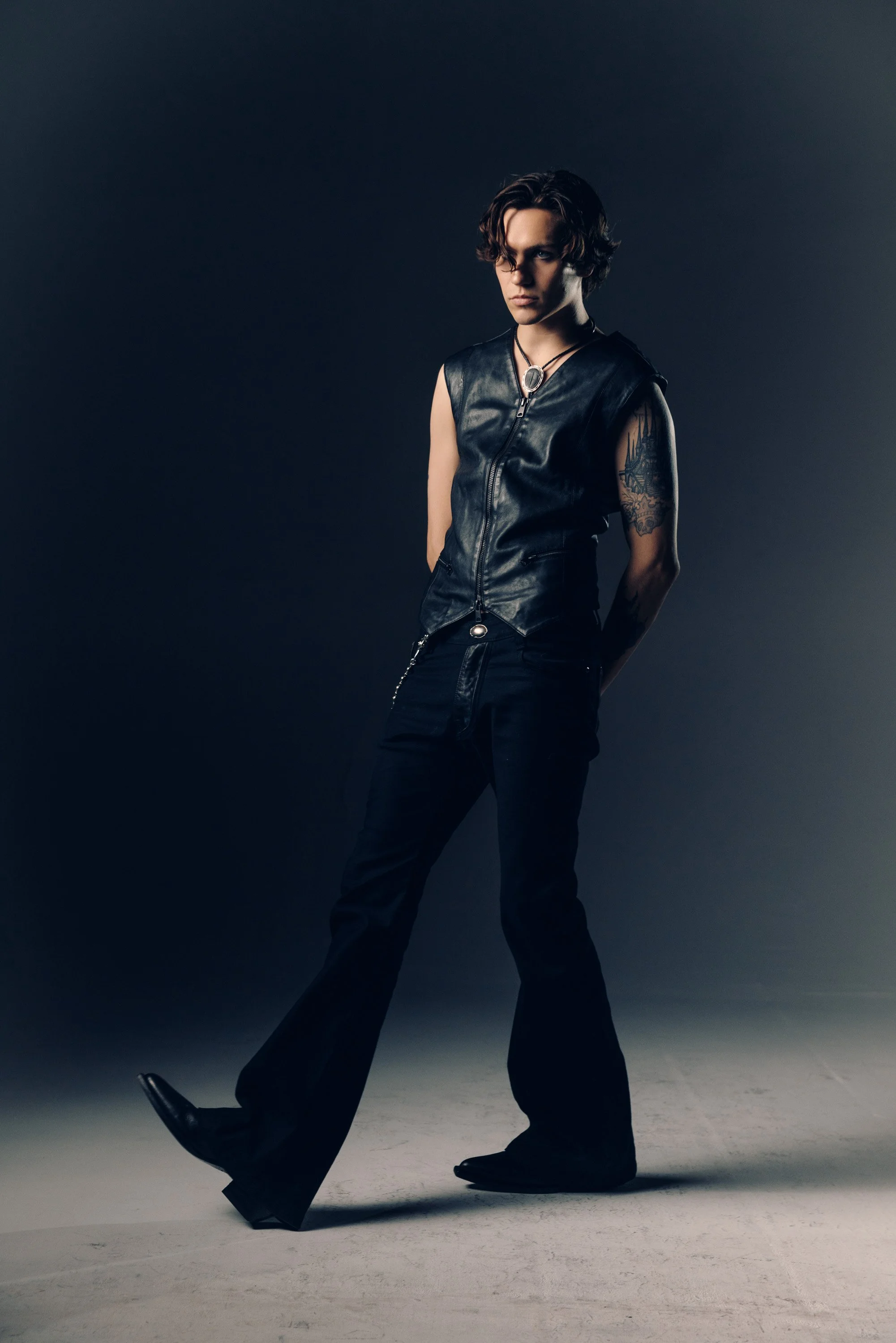 A person with curly brown hair and tattoos on their arm, dressed in a black leather vest and black pants, stands with their hands behind their back against a dark background, looking serious.