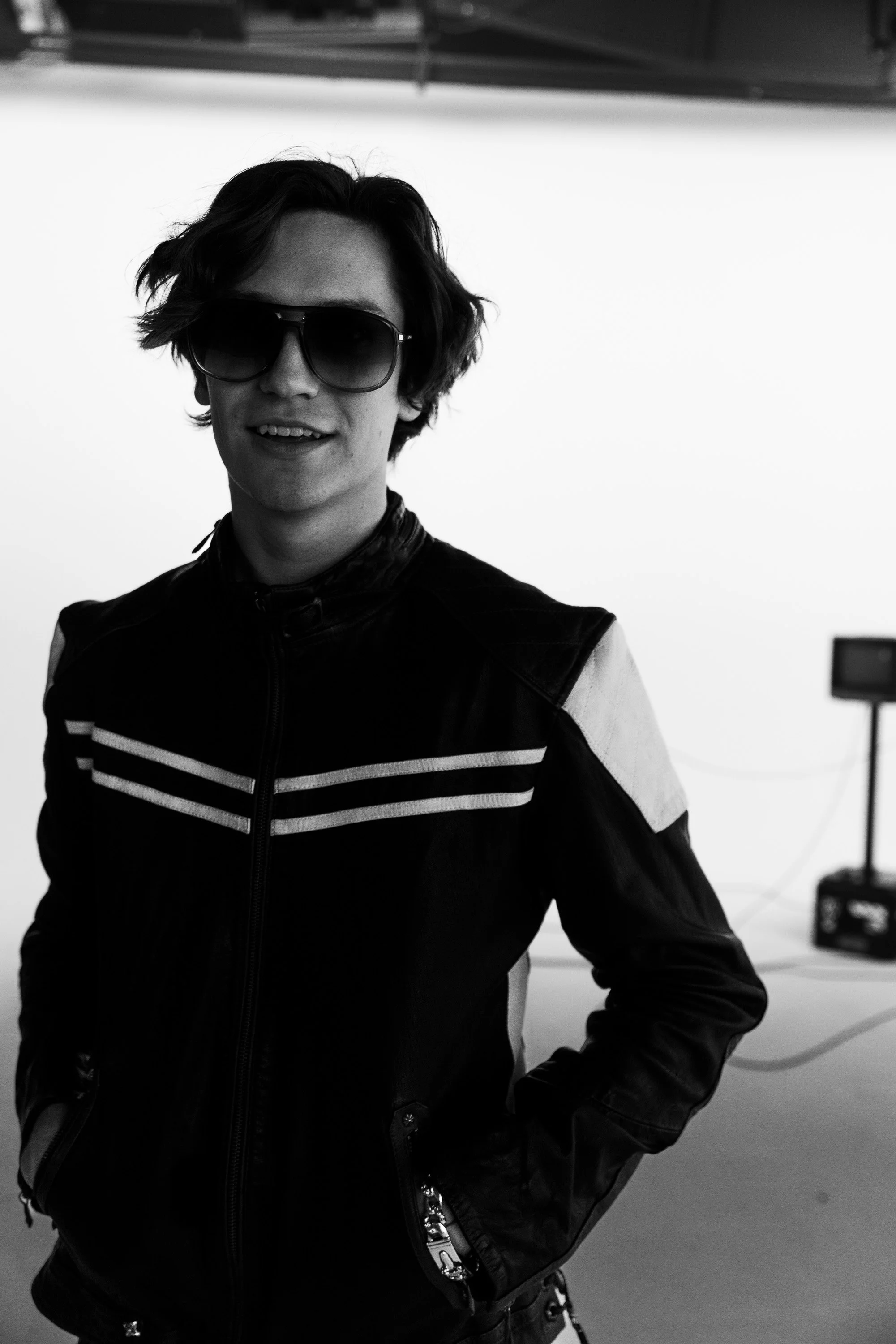 A person with wavy dark hair wearing large sunglasses, a black jacket with white stripes, smiling, and standing indoors.