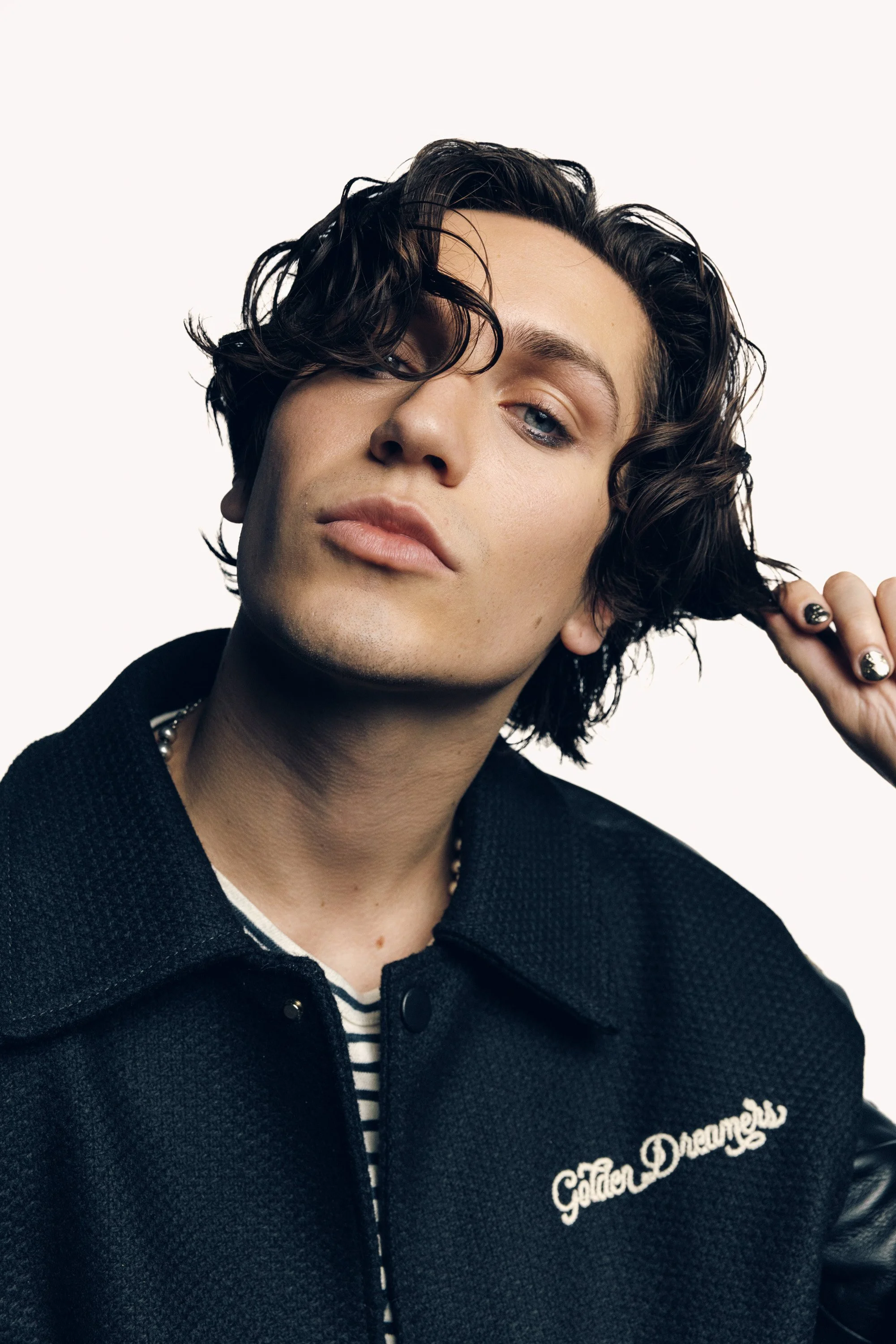 A close-up of a person with curly, dark hair, holding a lock of their hair, wearing a navy blue jacket with 'GoldenDreamers' embroidered on it, and a striped shirt underneath, in front of a plain white background.