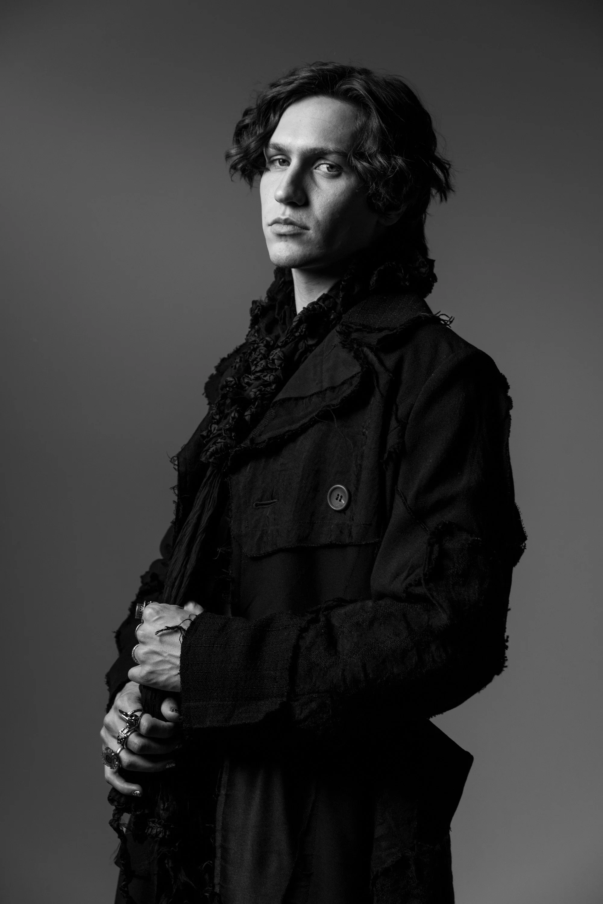 Black and white portrait of a young man with wavy hair, wearing layered dark clothing with rings on fingers, posing against a plain background.