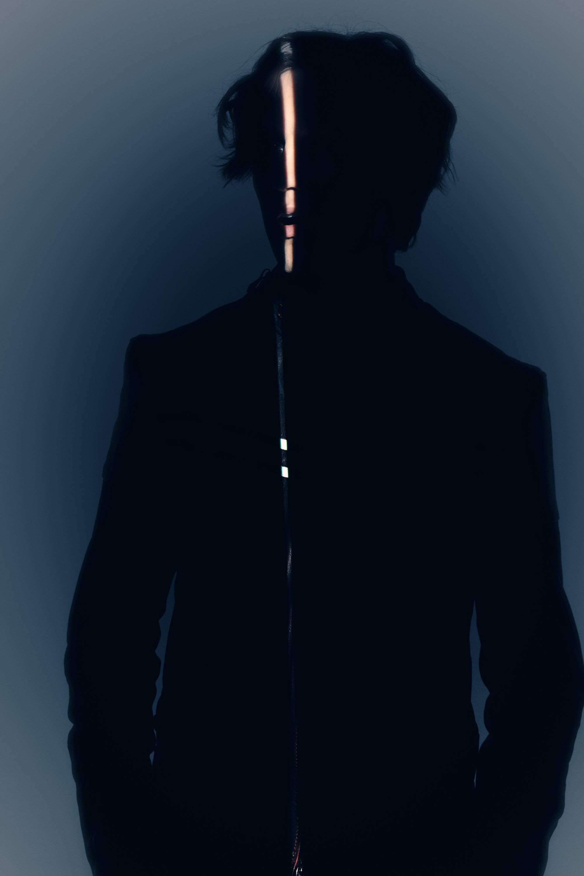 A person standing in front of a dark background with a vertical line of light running down their face, wearing a black jacket.