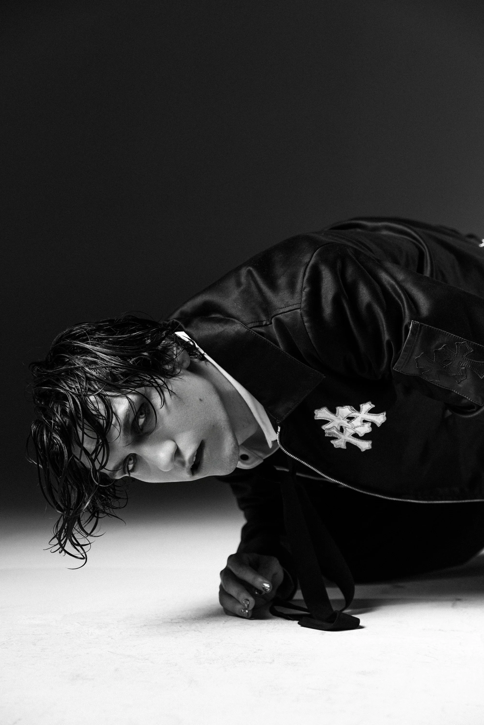 A person with wet, wavy hair wearing a dark leather jacket and a white shirt, kneeling on the floor and holding a martial arts black belt in their hand, against a plain dark background.