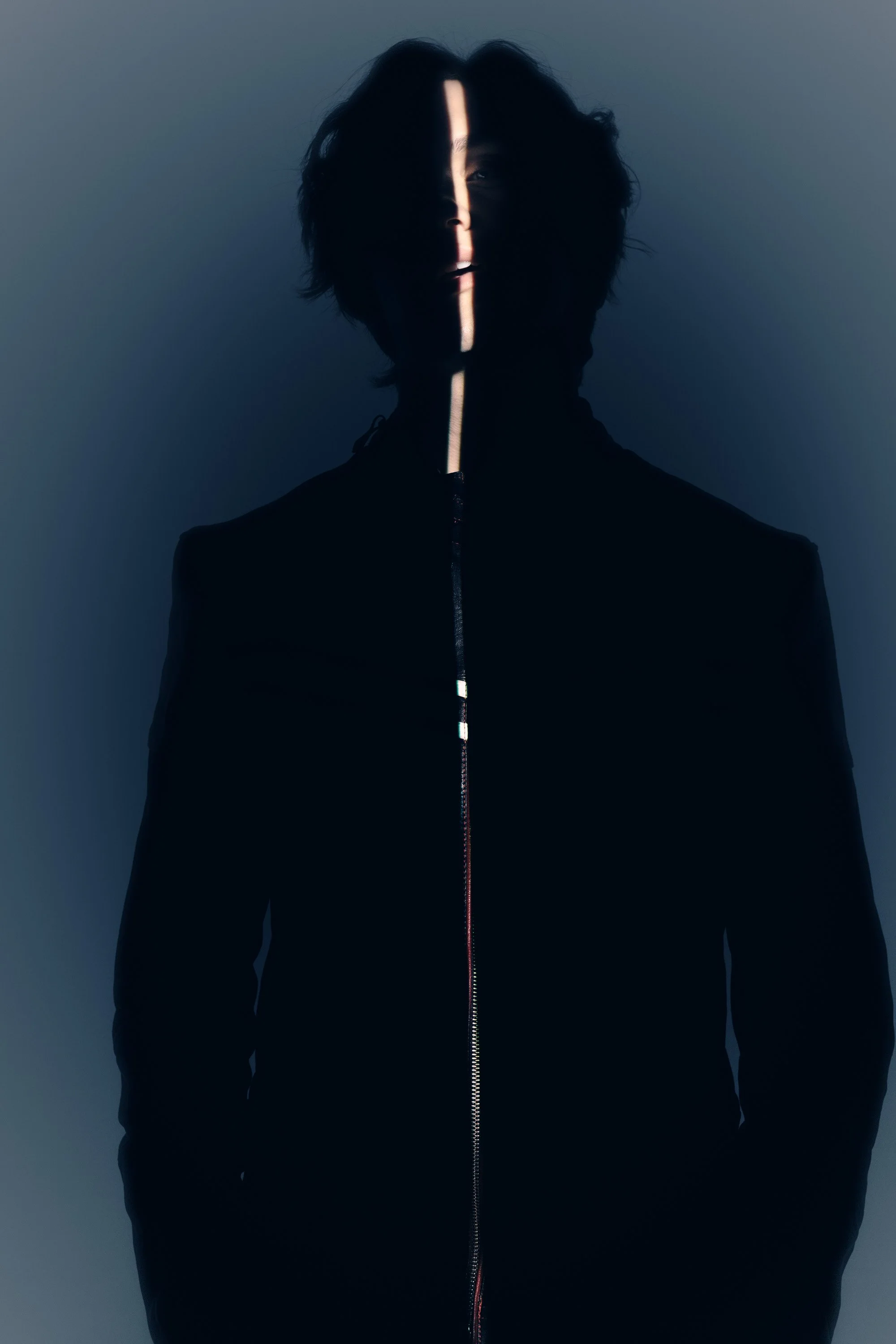 A person with medium-length black hair wearing a black jacket with a zipper stands against a gray background. A thin beam of light runs vertically down the person's face and jacket.