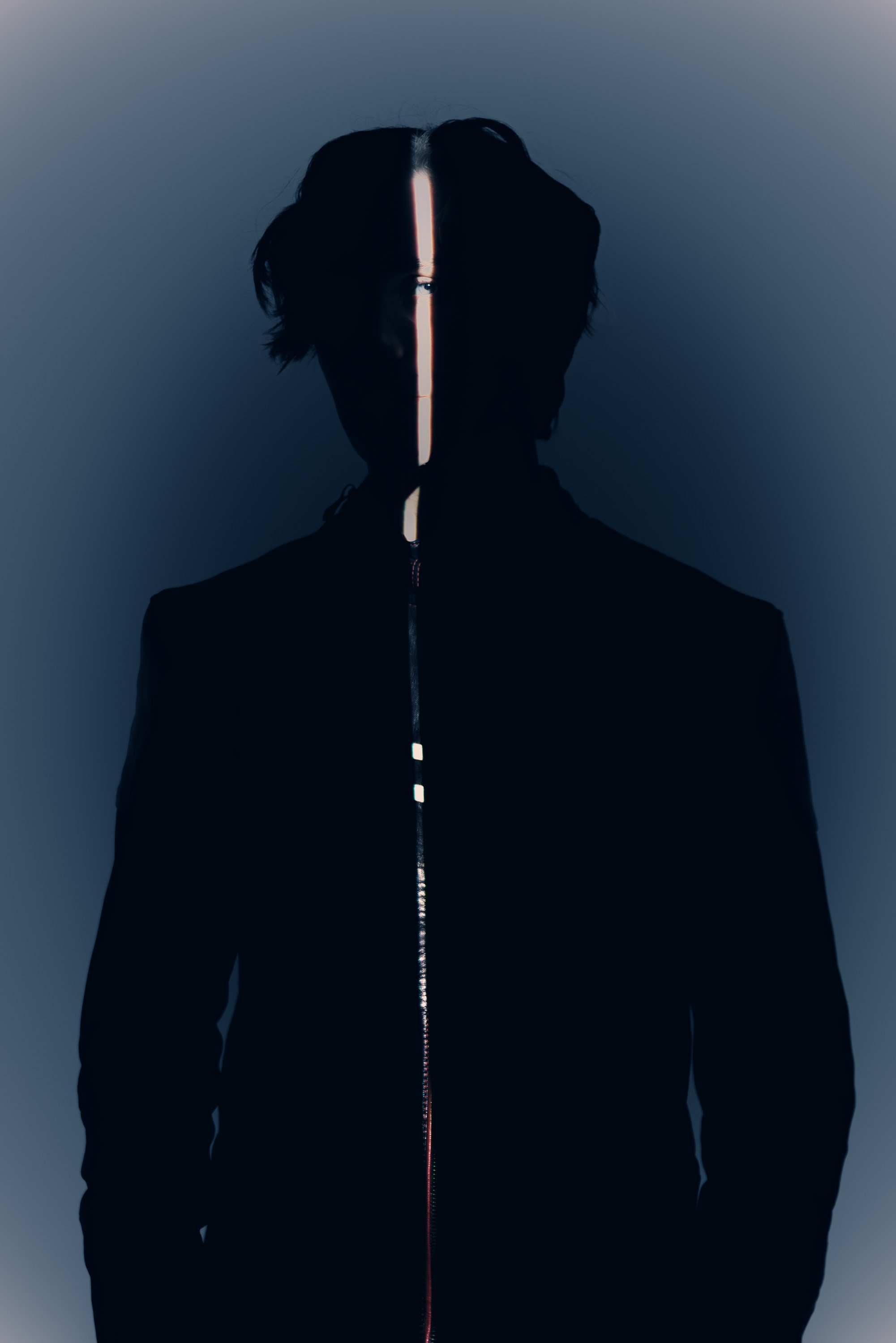 Silhouette of a person with dark hair, wearing a zip-up jacket, with a vertical neon light strip running down the middle of their face and body against a dark background.