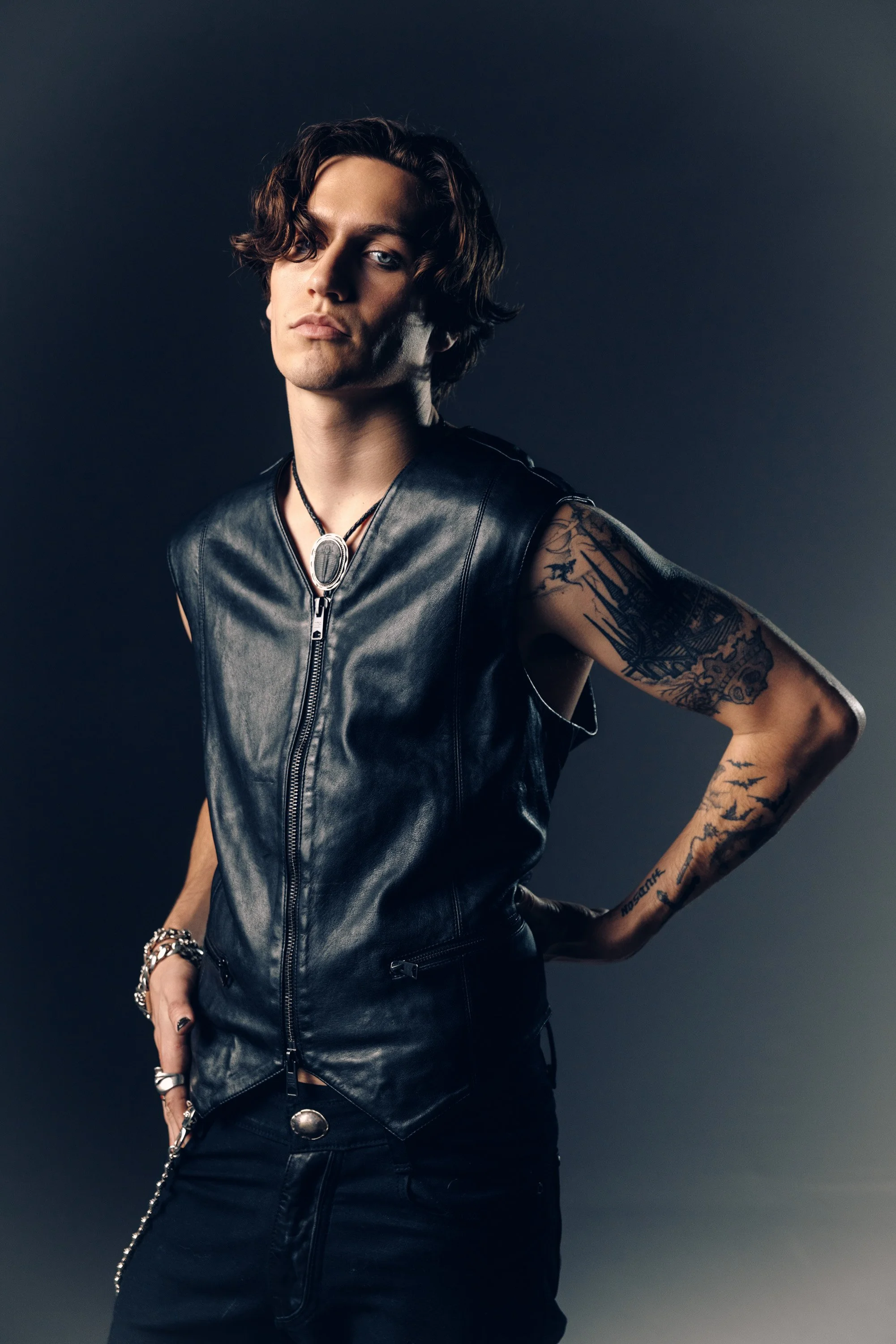 A person with wavy brown hair, wearing a black leather sleeveless vest, a silver pendant necklace, tattoos on their right arm, and multiple rings and bracelets, stands against a dark background with a confident pose.