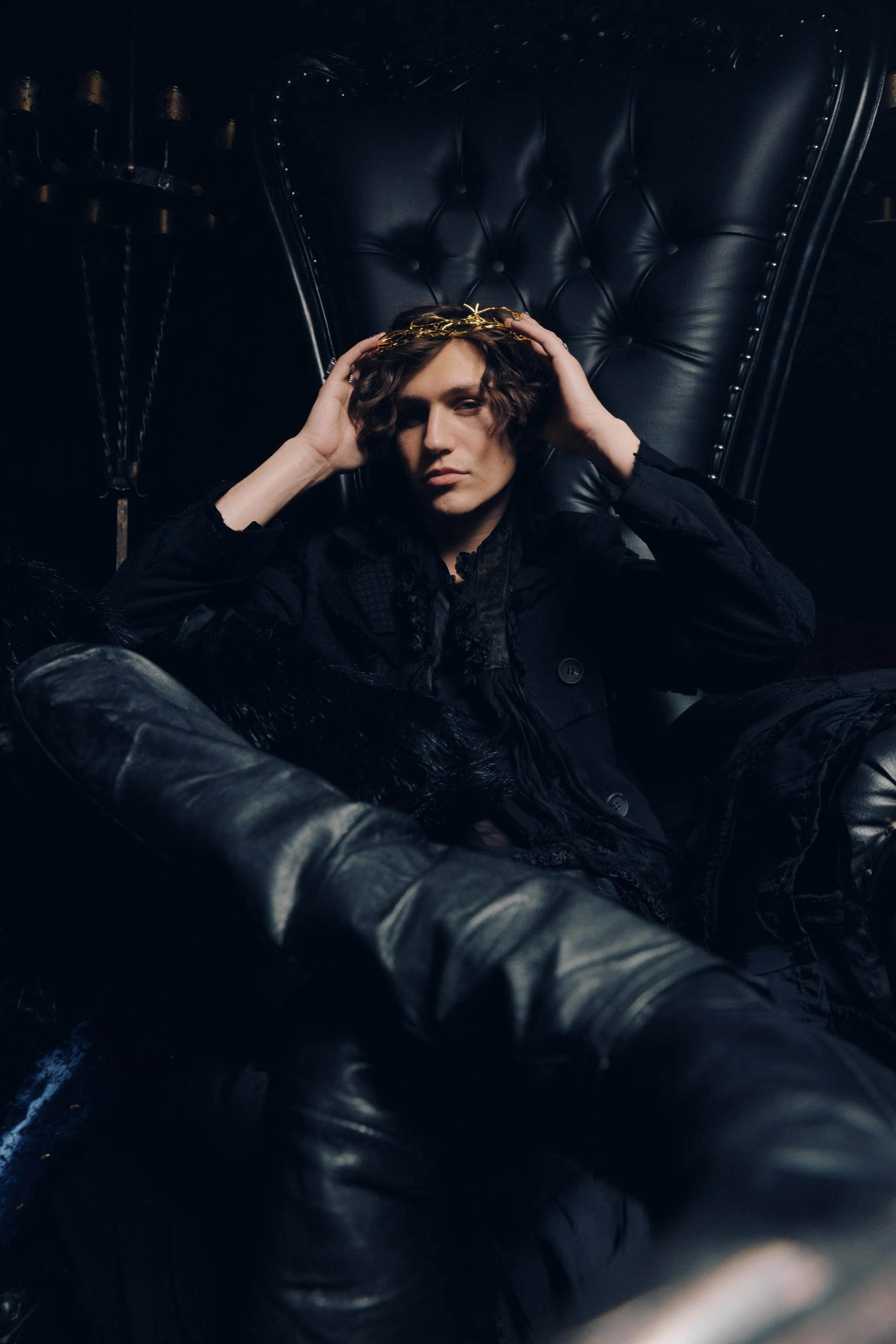 A person with curly hair sitting on a black leather chair, wearing a black jacket, and holding a gold crown on their head, in a dark room with a black background.