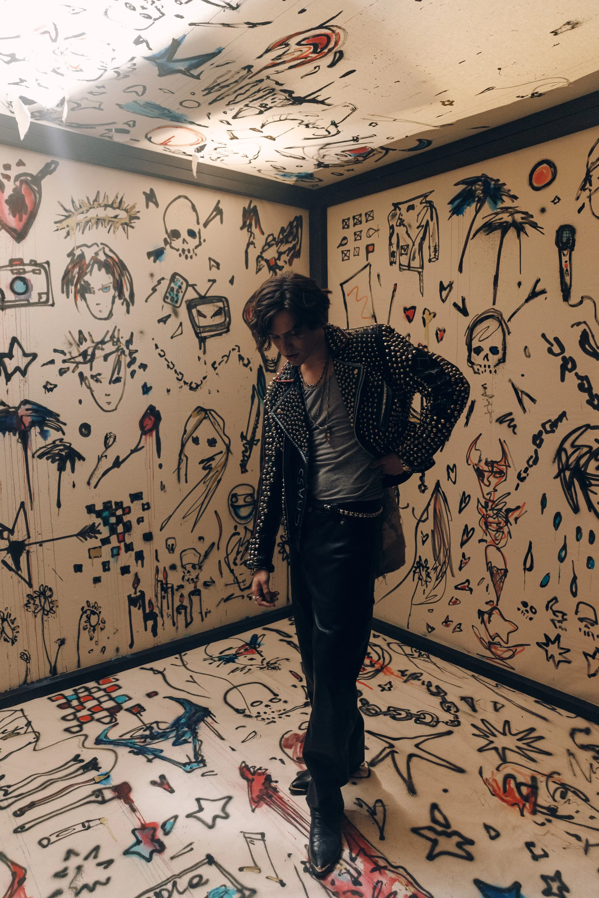 A person with wavy dark hair wearing a black leather jacket with metal studs, black leather pants, and black shoes standing in a room with walls, ceiling, and floor covered in colorful, cartoon-style doodles and sketches.