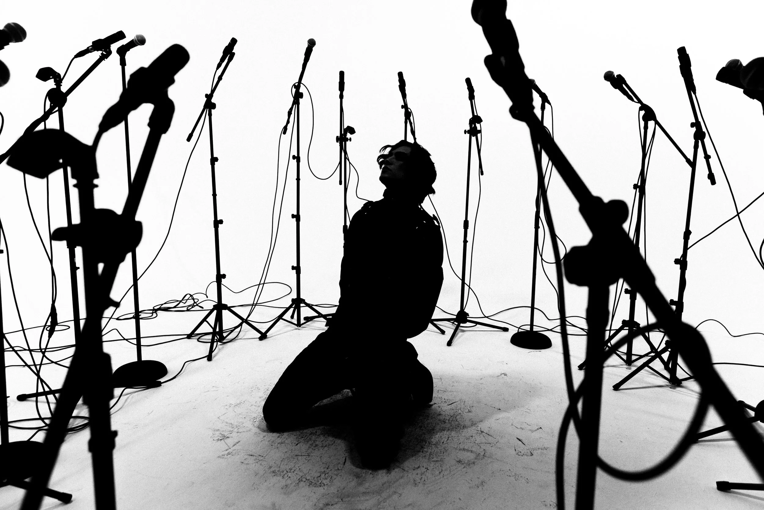 Black silhouette of a person kneeling on the floor surrounded by multiple microphone stands, microphones, and cables, against a white background.