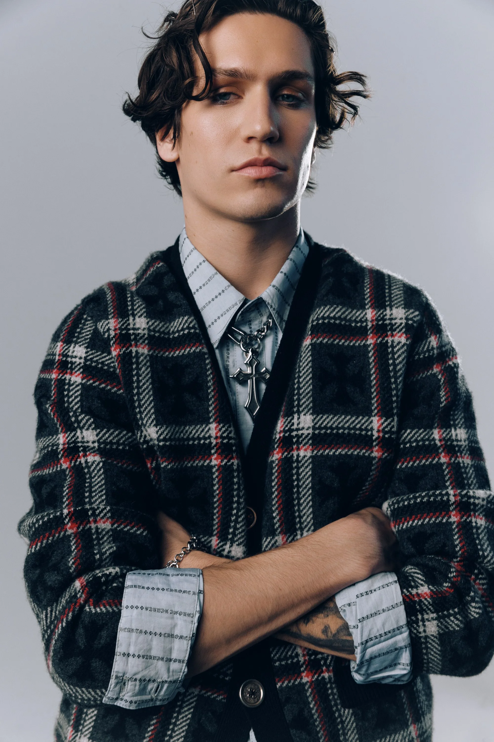 A person with wavy brown hair, wearing a plaid jacket over a patterned shirt, is standing with arms crossed against a neutral background.