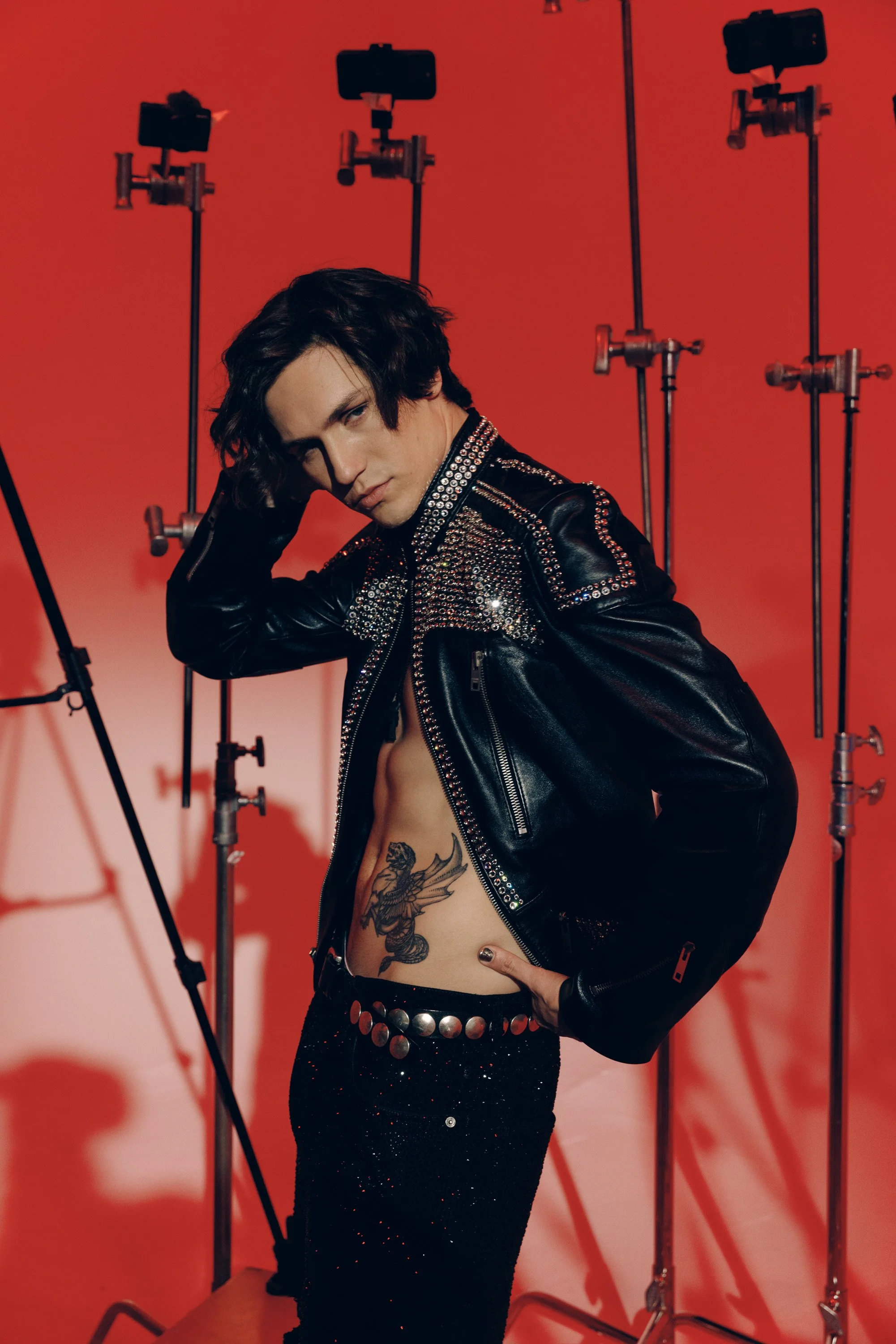 Person with long dark hair and tattoos on torso, wearing a black leather jacket with silver studs and sparkly black pants, poses against a red background with photography equipment.