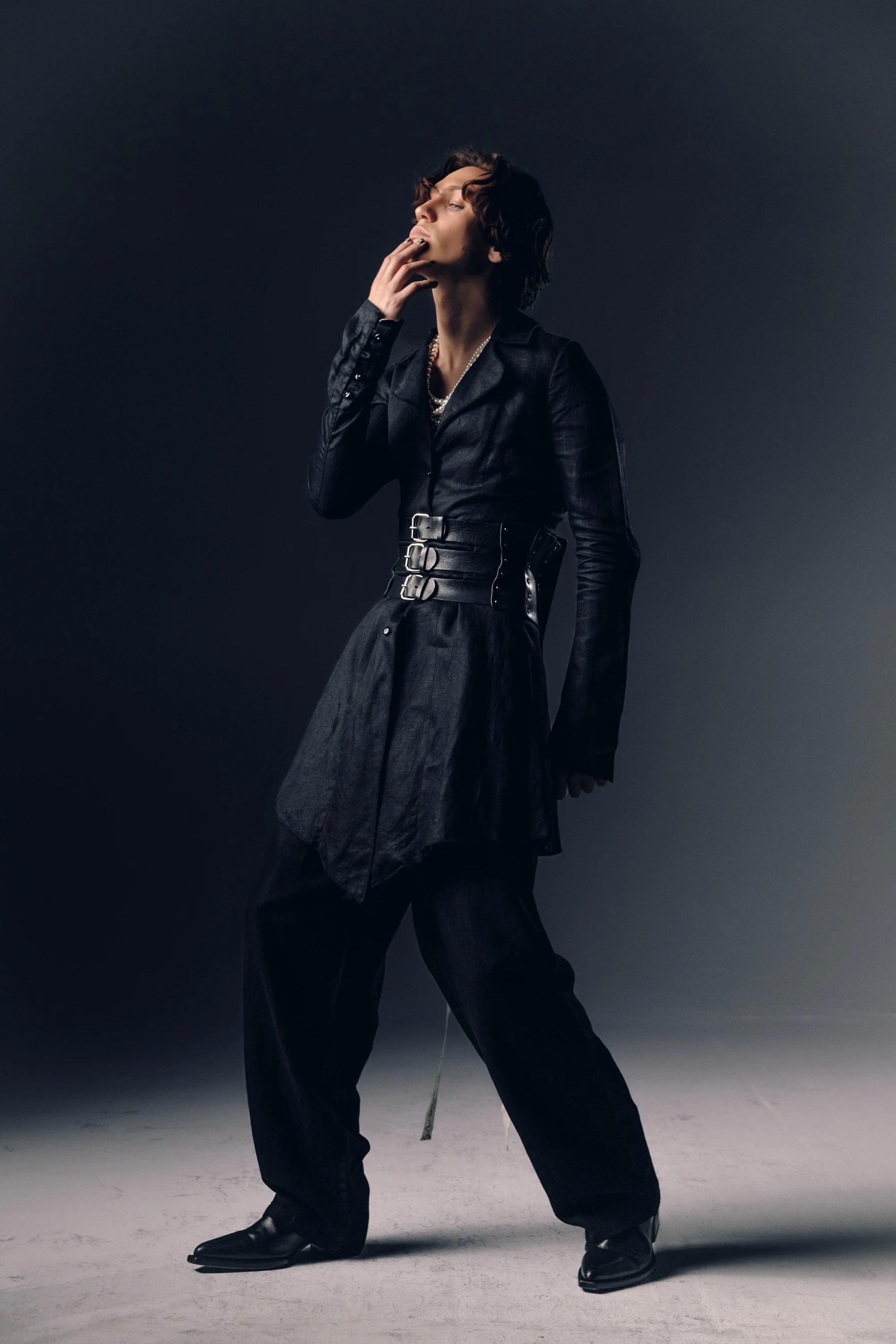 A person with short, curly hair wearing a dark, stylish outfit, including a belted trench coat, oversized pants, and boots, posing against a neutral background with dramatic lighting.
