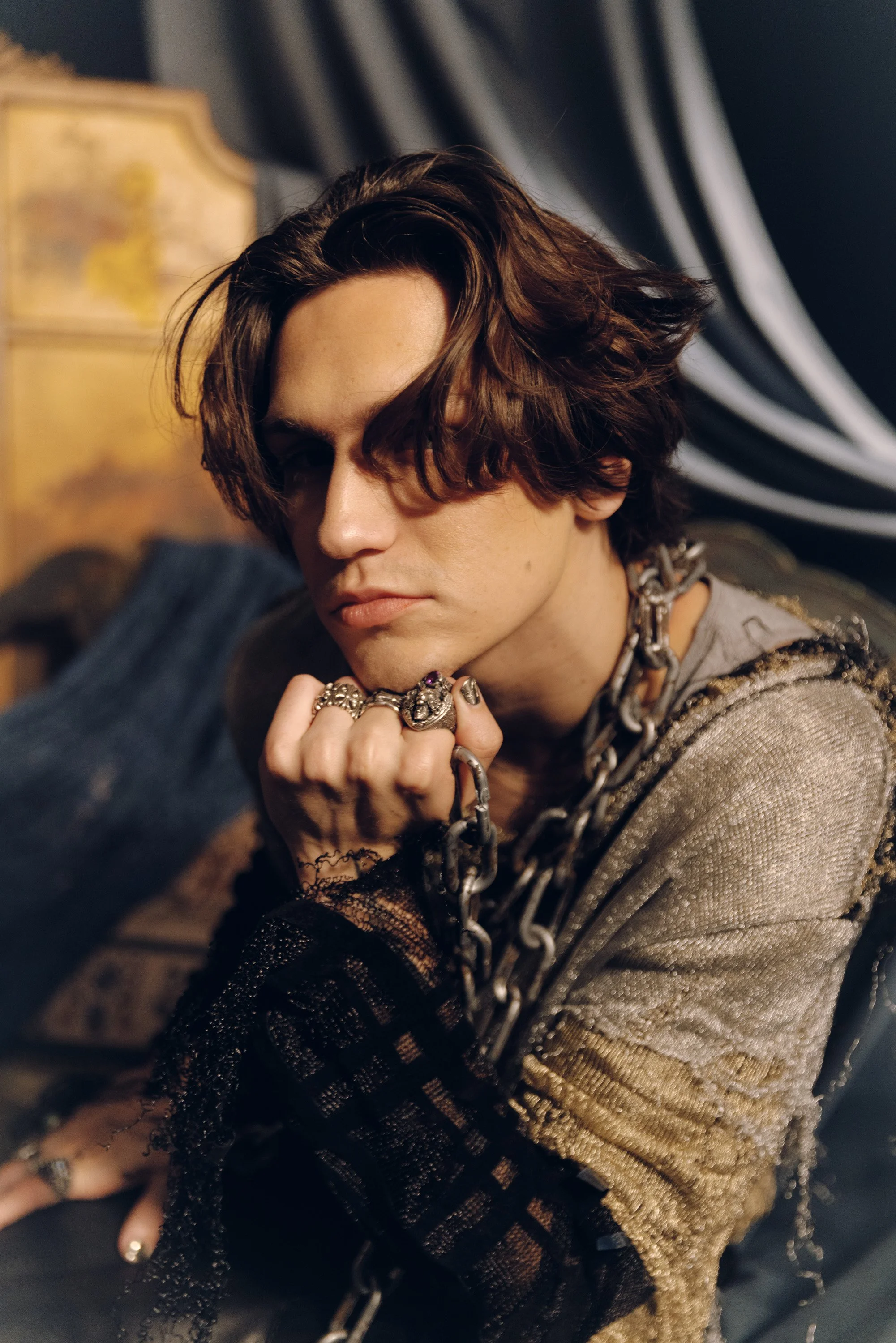 Close-up of a young person with medium-length wavy brown hair, wearing a silver chain necklace and multiple silver rings, with their right hand resting on their chin. The person is dressed in a gray and black striped outfit, with a black lace glove o