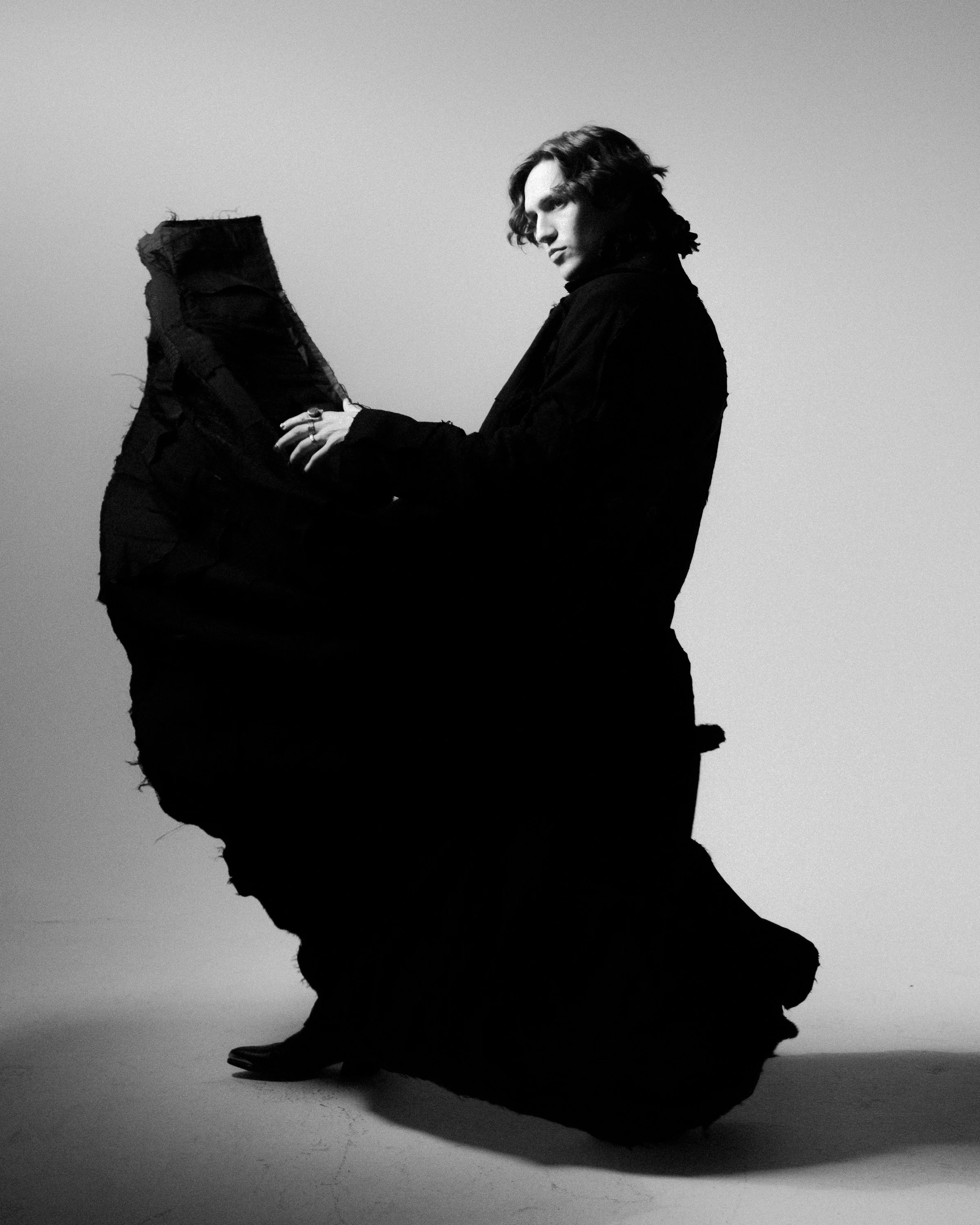 Black and white photo of a young man with curly hair, dressed in dark clothing, standing sideways and looking down, with a shadow cast on the floor.