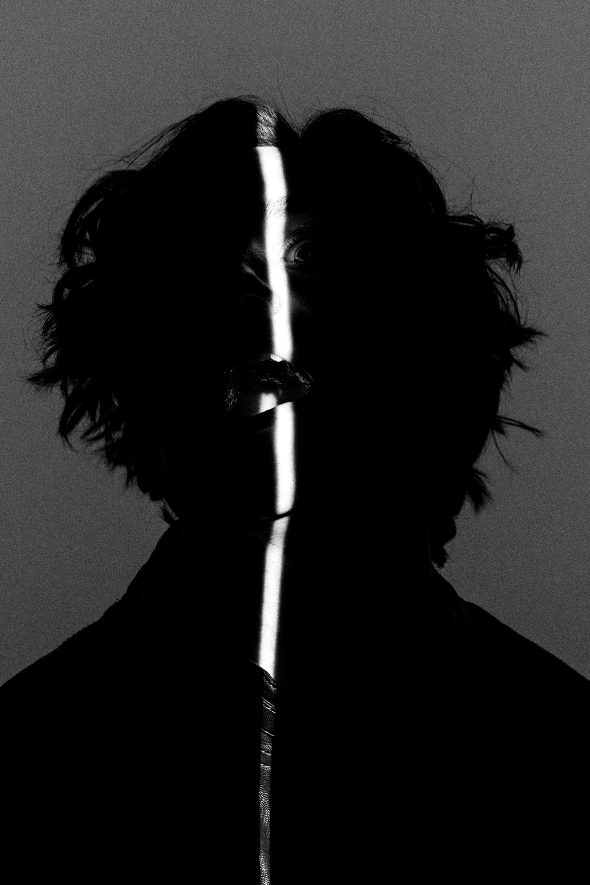 Black and white photograph of a person's face with a vertical line of light dividing the face into two halves, with the person's eye visible on the left side of the line.