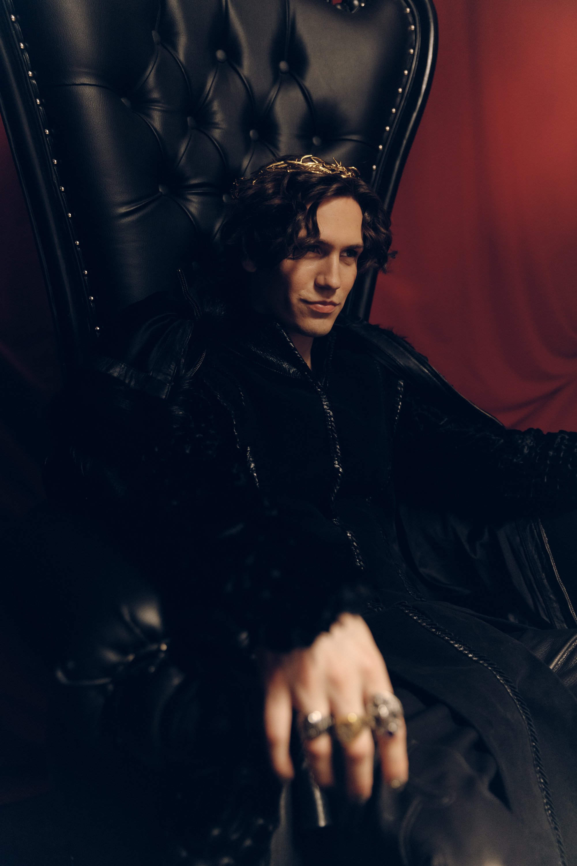 Person with dark, curly hair sitting on a black leather chair with a high back and nailhead trim, wearing a black leather jacket, against a red wall.