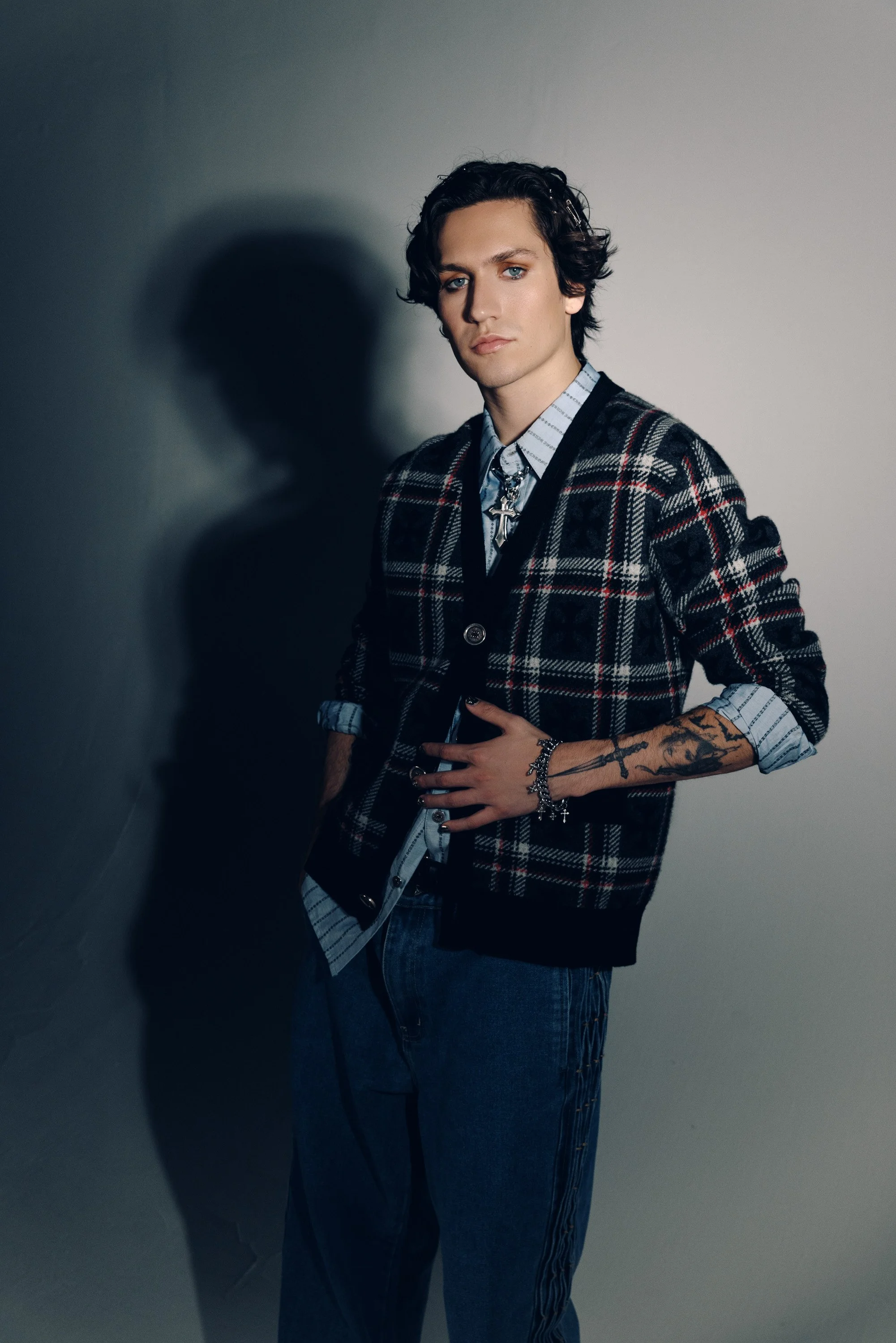 A young man with wavy dark hair and tattoos on his left arm, wearing a plaid blazer over a button-up shirt and jeans, stands against a plain wall with shadows cast on it.