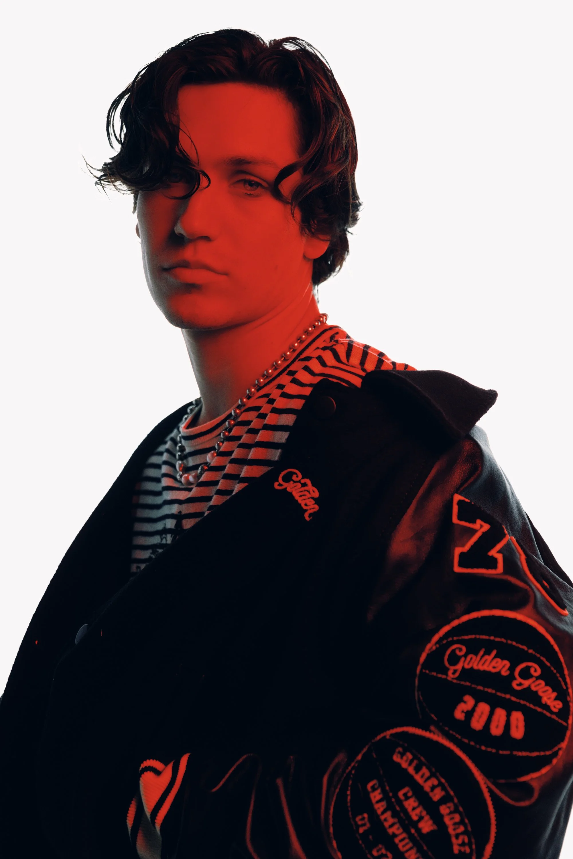 A young man with wavy dark hair wearing a striped shirt, a black jacket with red embroidery and patches, and a pearl necklace, against a plain light background.