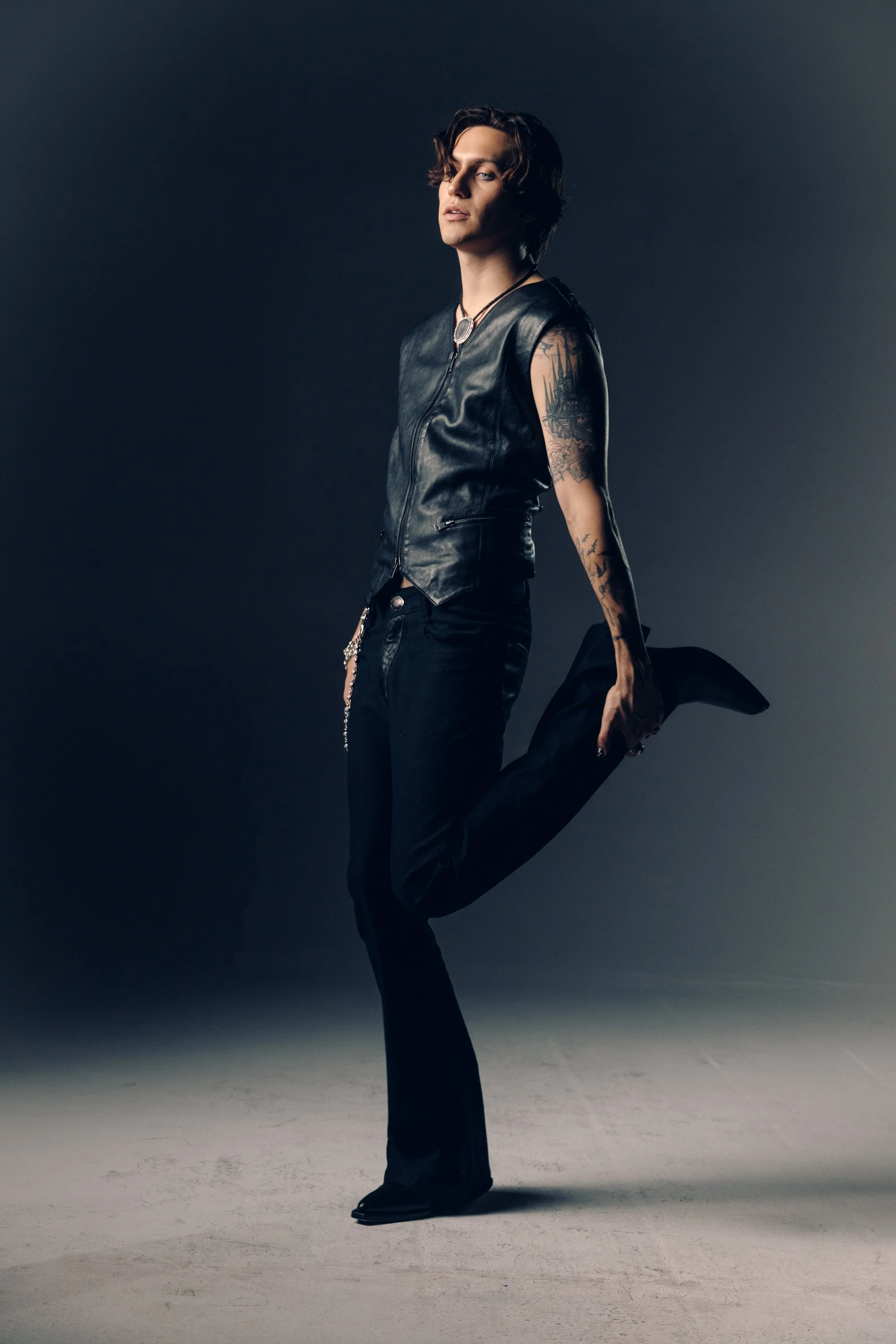 Man with wavy brown hair wearing black leather vest, black pants, black boots, and a necklace with a pendant, standing against a dark gradient background.