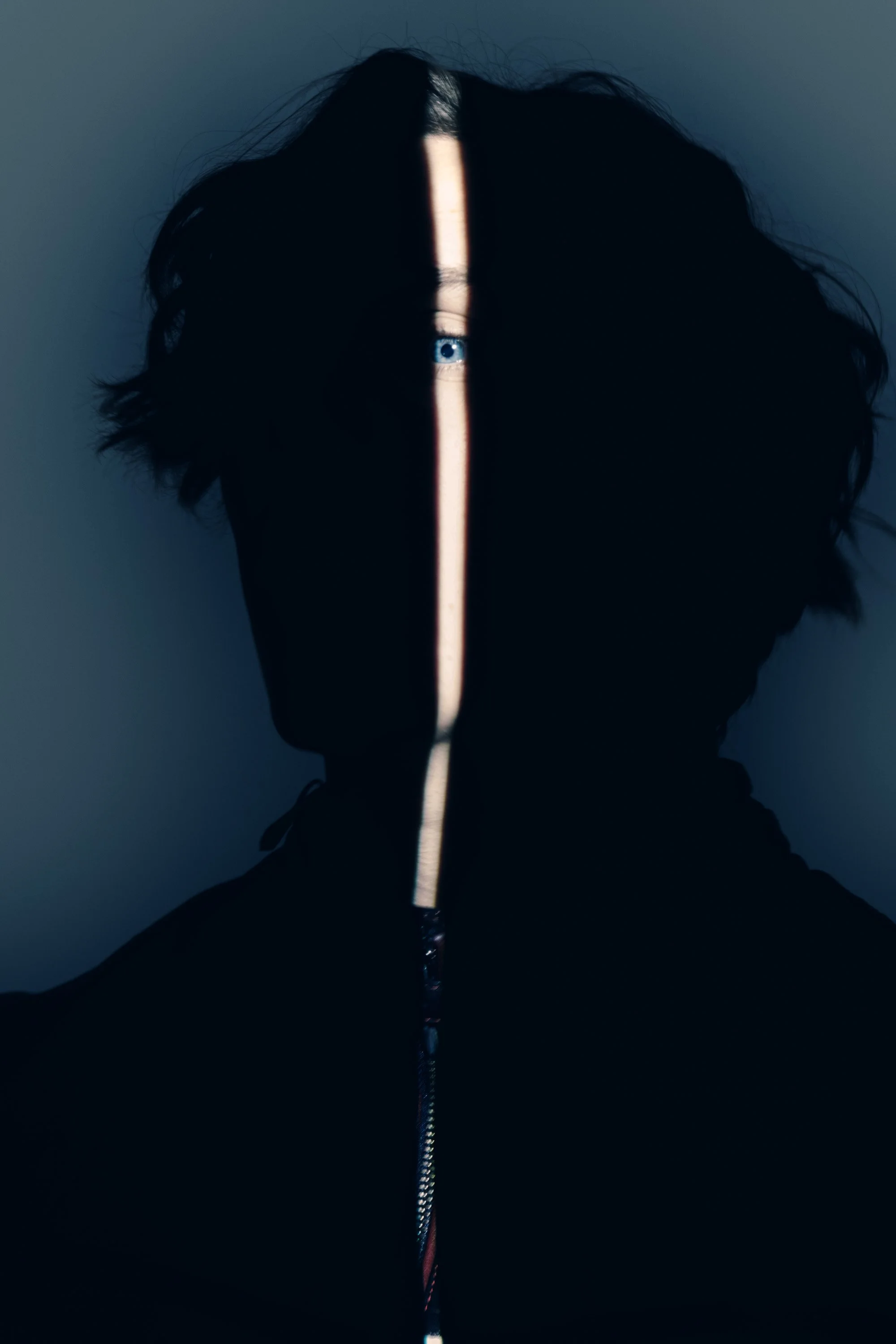 A person's silhouette with wild, curly hair, illuminated by a narrow vertical beam of light running from the top to the bottom of the image, revealing a single blue eye.