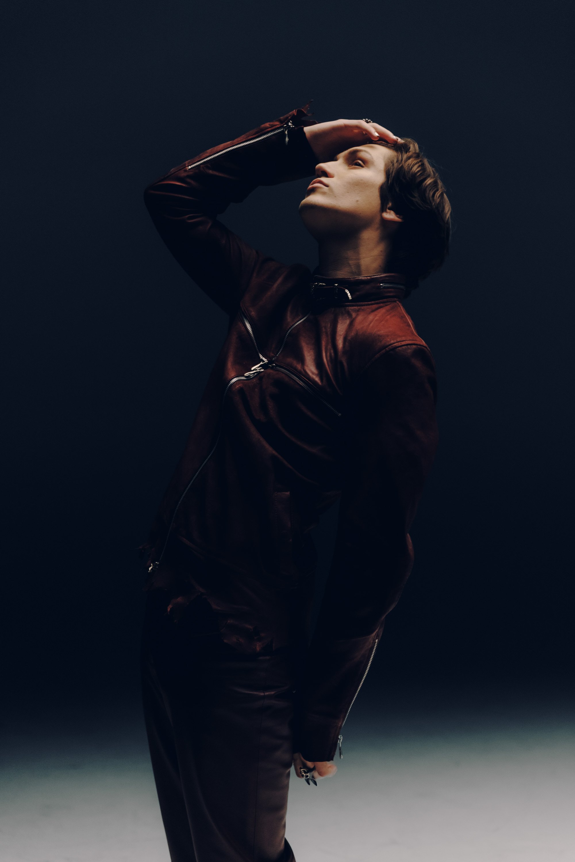 Person with short dark hair wearing a red leather jacket and dark pants, posing against a solid dark background with their hand on their forehead.