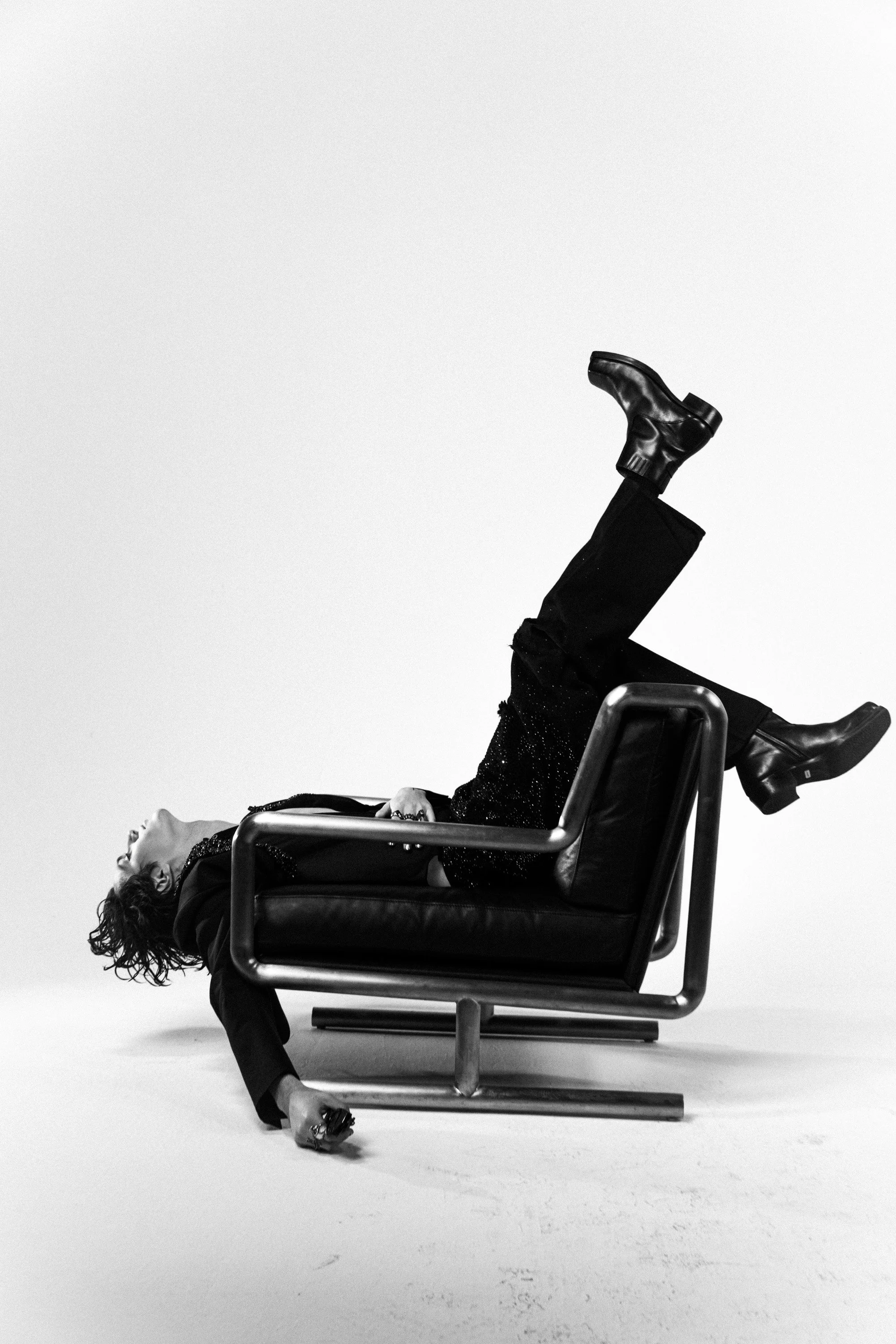 Black and white photo of a person lying on a chair with their legs elevated as if in a playful pose, with curly hair and stylish clothing.