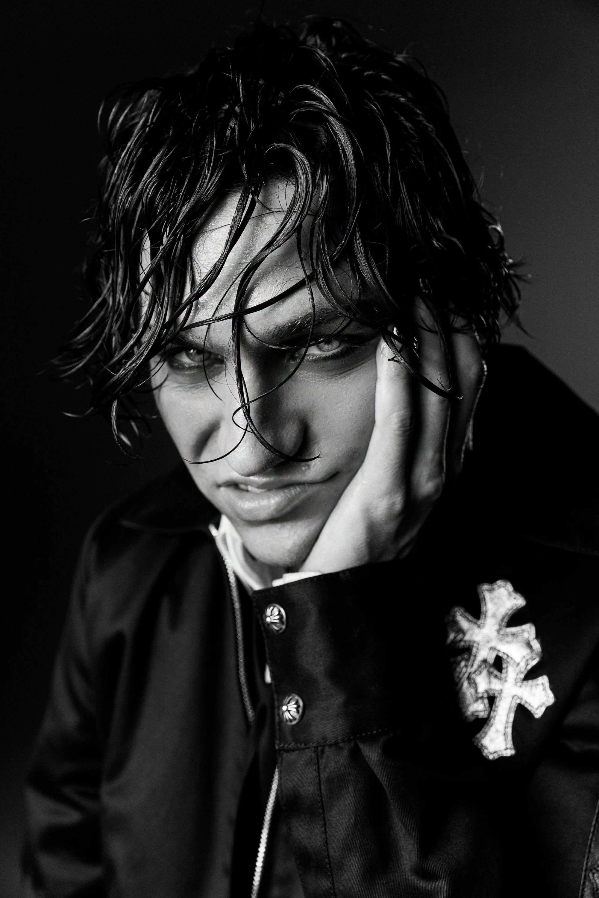 Black and white photo of a person with wet, wavy hair covering part of their face, wearing a dark jacket with a large cross emblem on the chest, and looking down with one hand near their face.