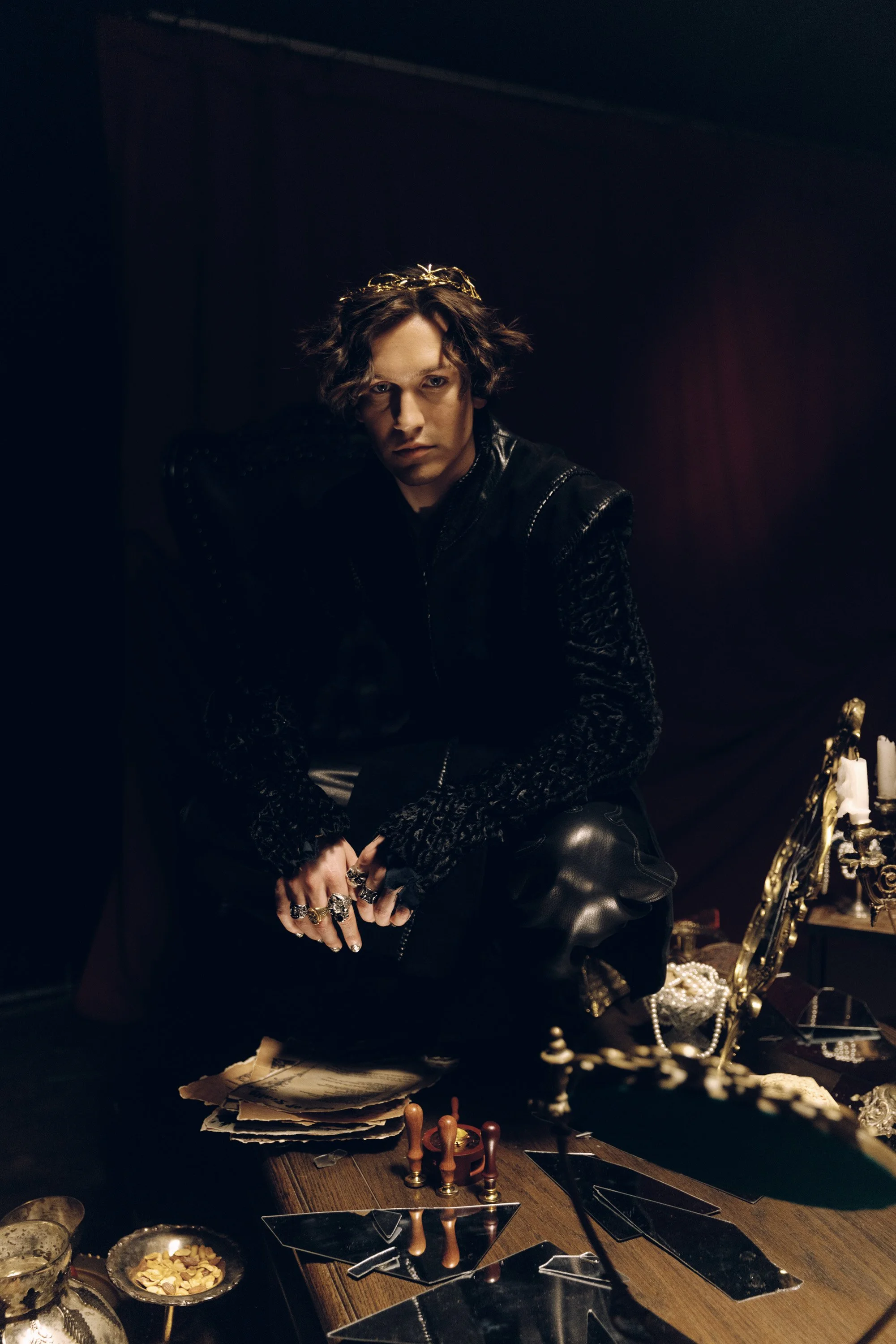A person with wavy hair wearing a black leather jacket and multiple rings, sitting at a cluttered table with jewelry and papers, with dark background and red lighting.