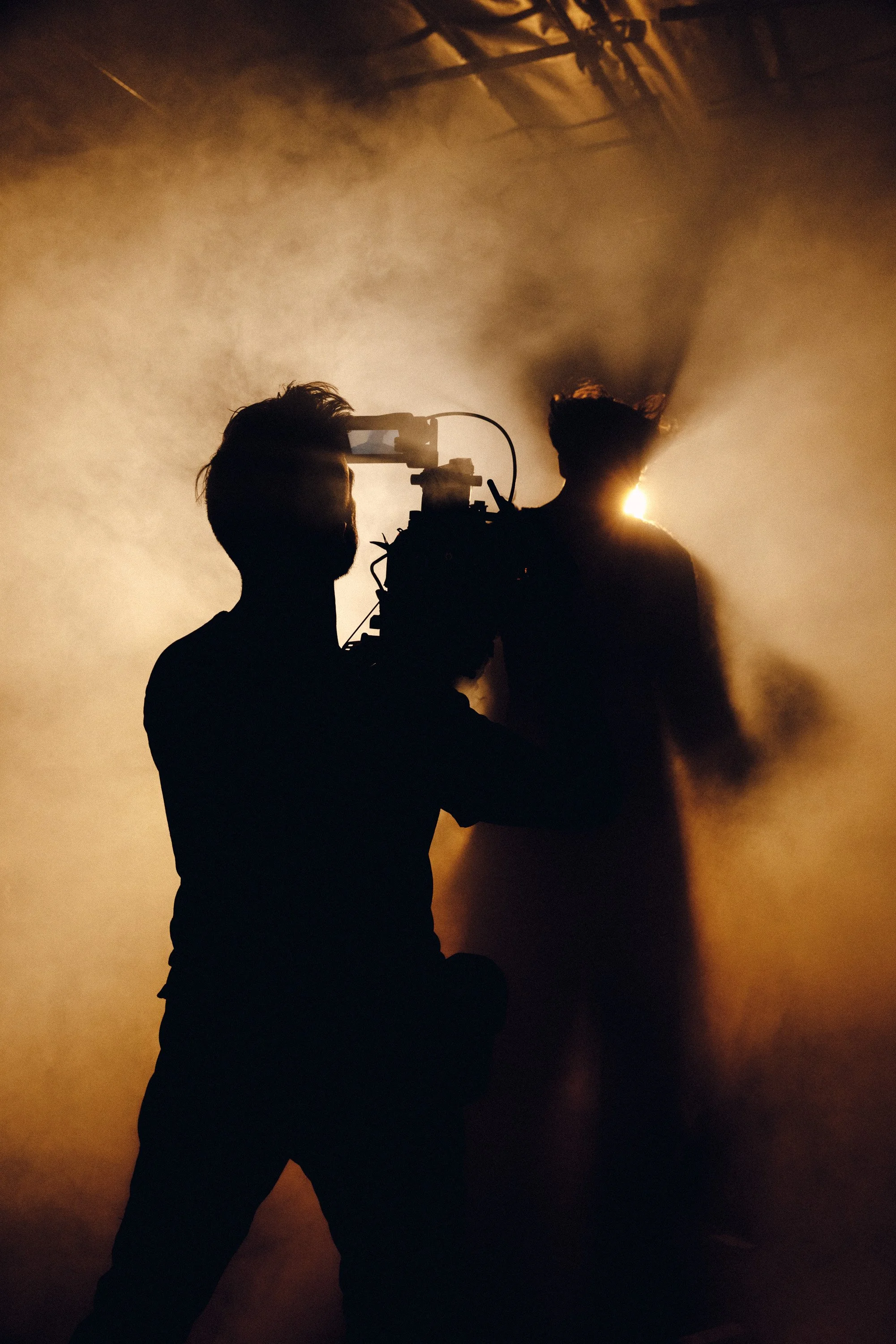 A silhouette of a person in a dark room, holding a camera or filming device, with a bright light shining behind them and smoke or fog creating a dramatic effect.